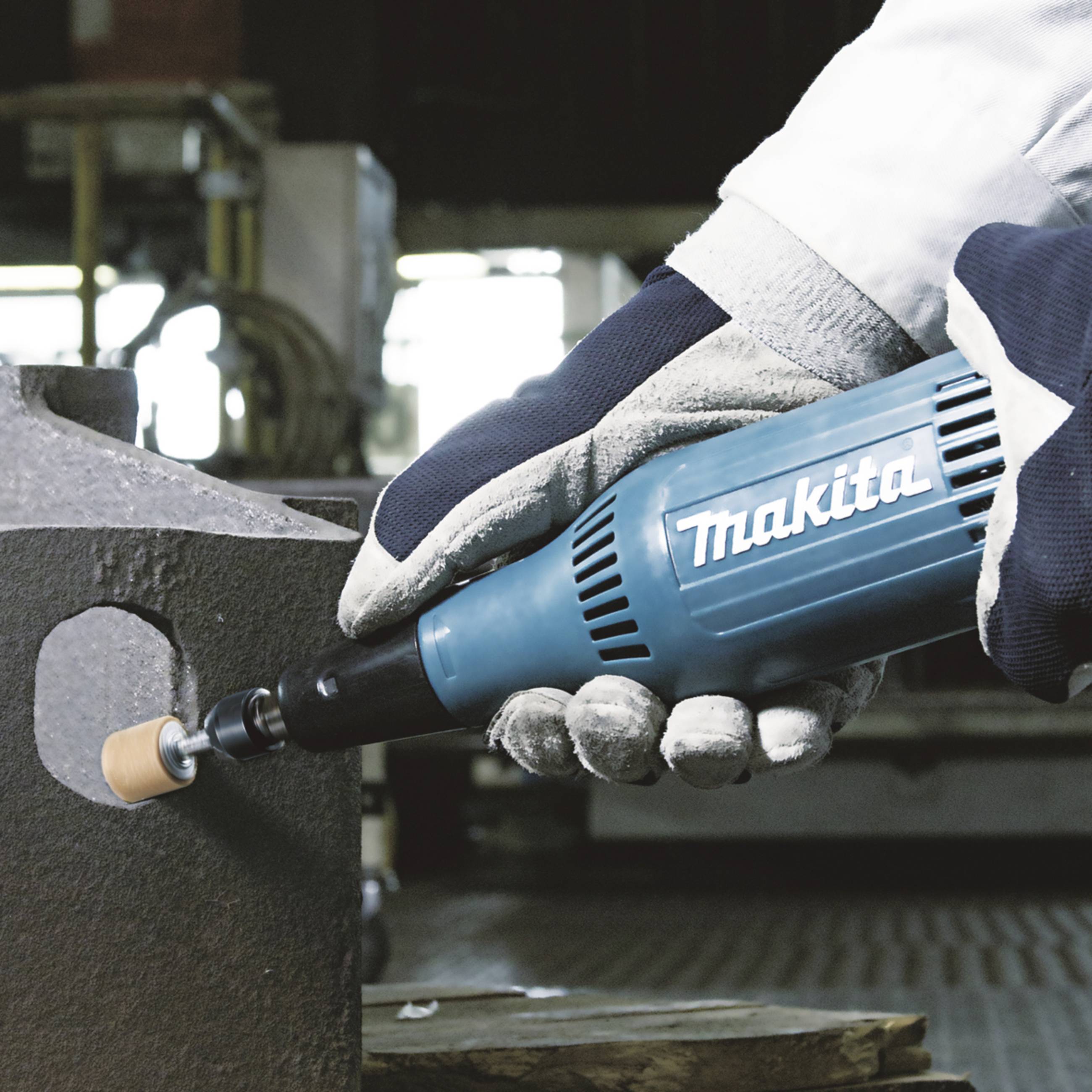 Travailleur portant des gants de protection qui meule du métal avec un outil Makita bleu ; environnement industriel en arrière-plan.