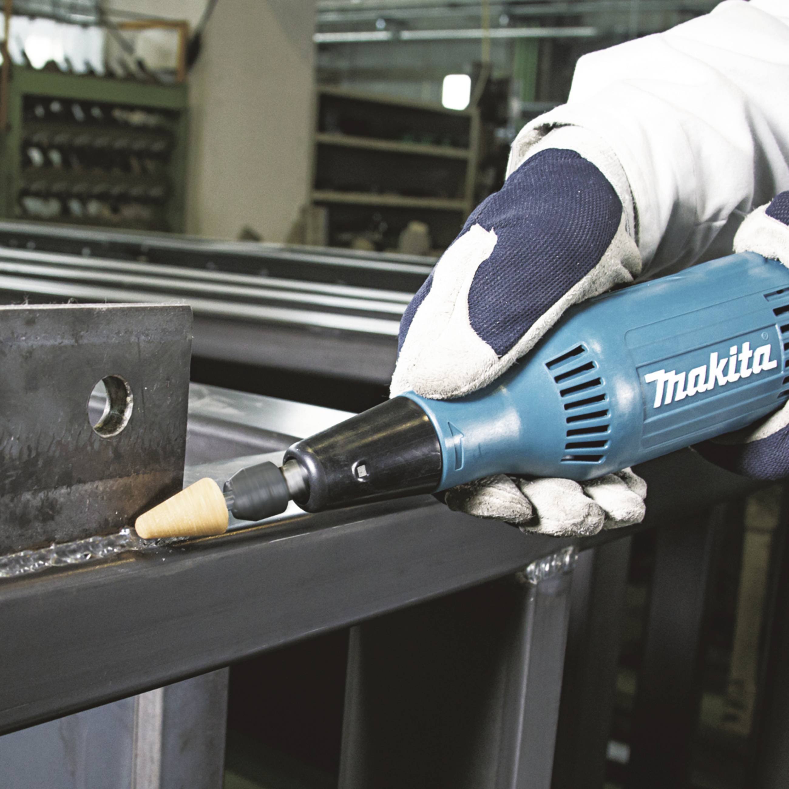 Une personne portant des gants de protection tient une meuleuse d'angle Makita et travaille une surface métallique dans un environnement d'atelier.