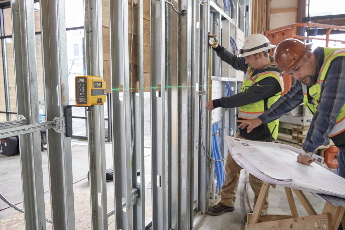 Des ouvriers sur un chantier vérifient des plans de construction devant une structure de cadre métallique. Un appareil de nivellement laser est installé sur le mur.
