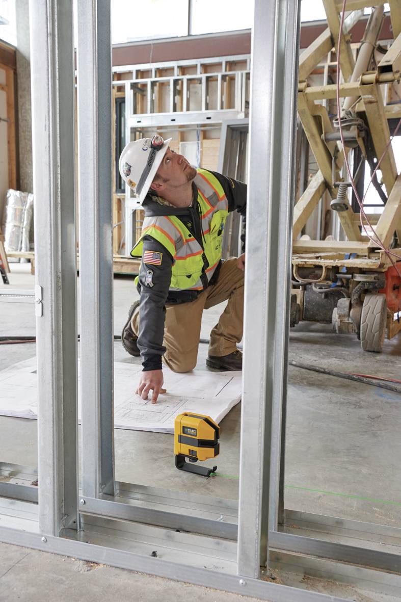 Un ouvrier portant un casque de chantier et un gilet de sécurité est agenouillé sur un chantier et vérifie des plans de construction à côté d'un appareil laser.