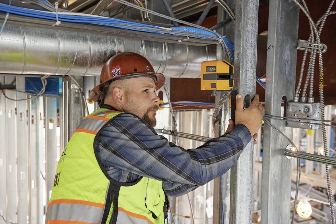 Un ouvrier en tenue de protection utilise un appareil de mesure dans un bâtiment en construction. Des câbles électriques et des tuyaux sont visibles.