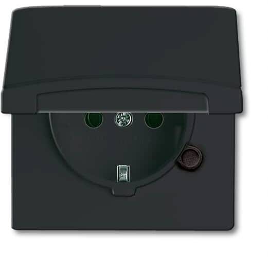 Une prise de courant avec terre, grise, à volet rabattable, montée sur un mur. Adaptée pour une utilisation en extérieur.