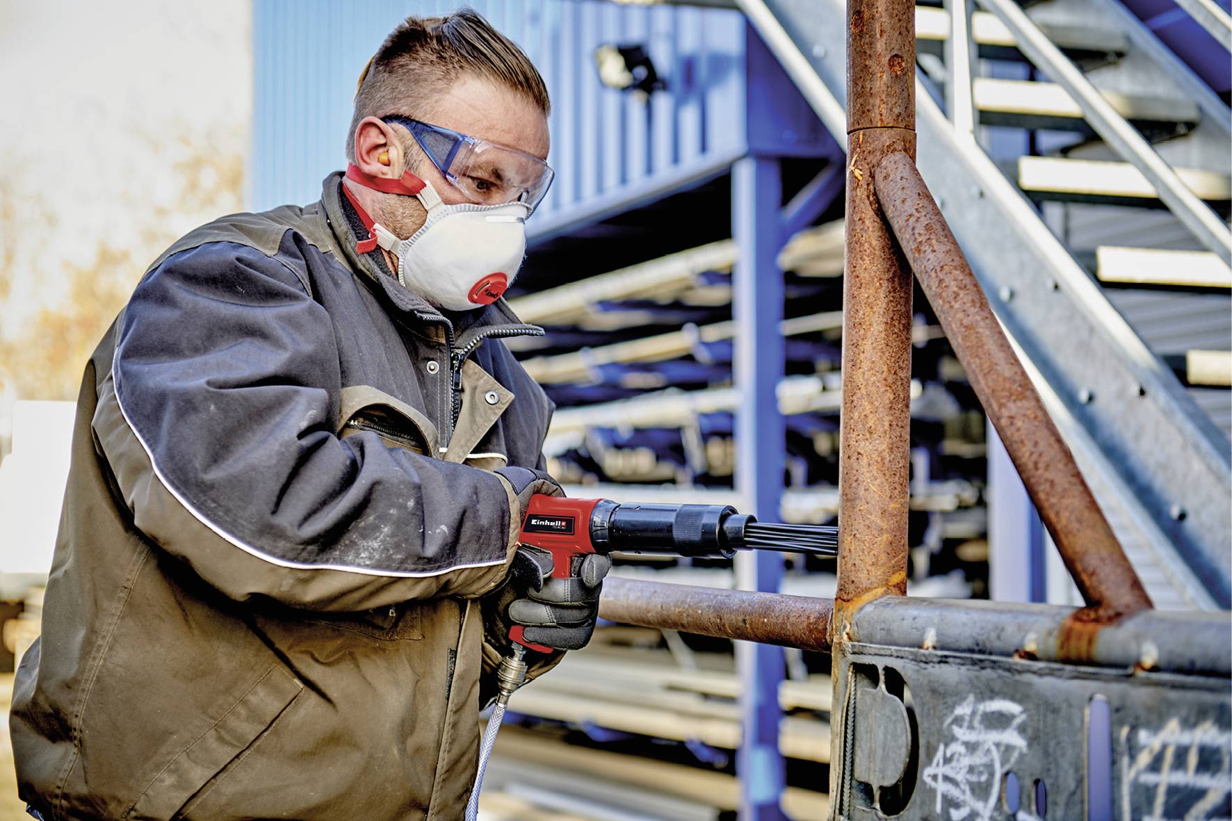 Un ouvrier porte un équipement de protection, des lunettes de sécurité et un masque respiratoire, alors qu'il travaille avec un outil électrique sur une rambarde métallique.