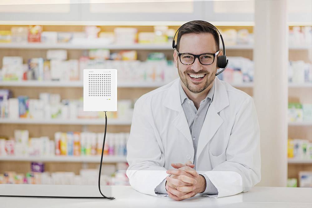 Un pharmacien souriant, portant un casque, se tient derrière un comptoir dans une pharmacie. Des étagères de médicaments sont visibles en arrière-plan.