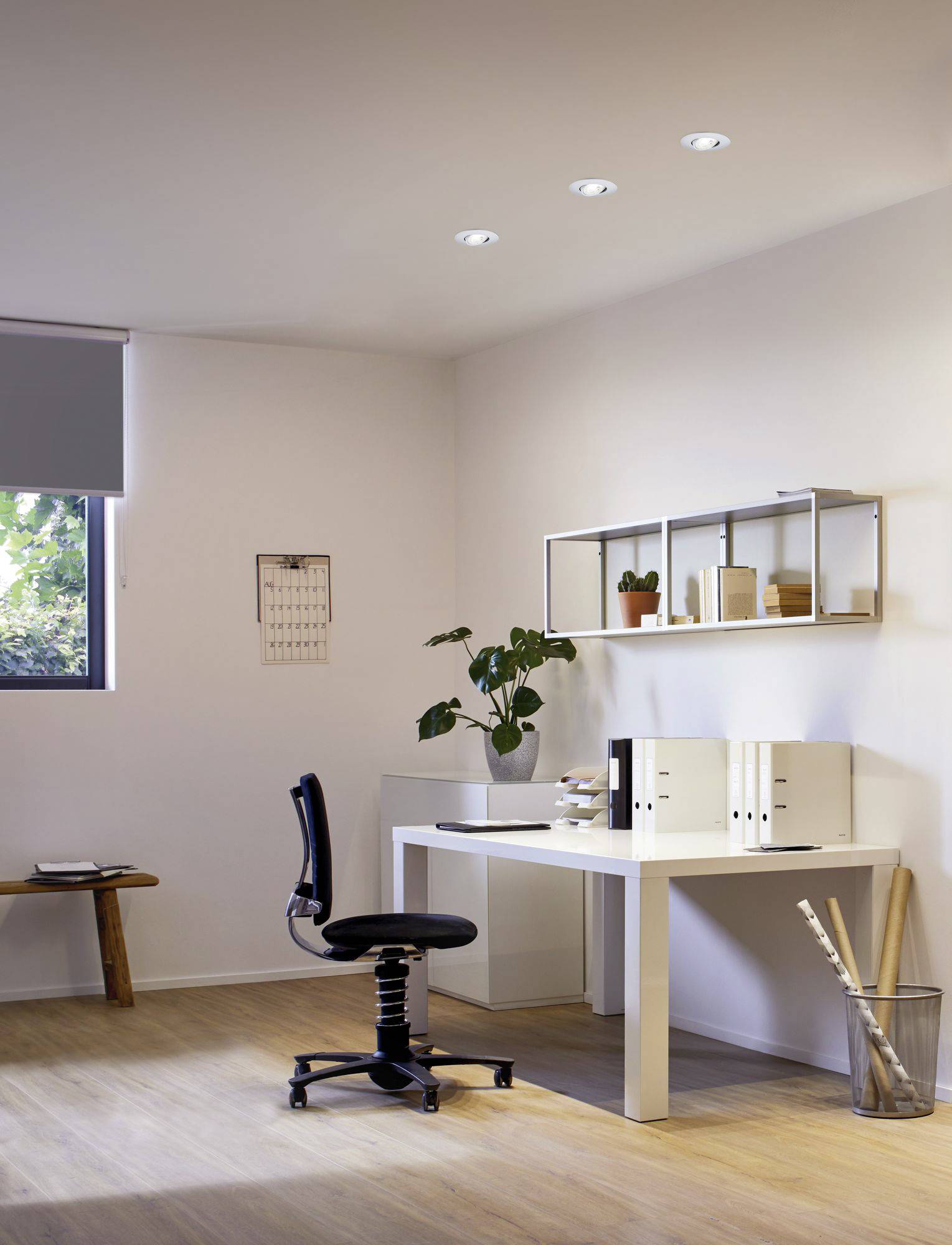 Un bureau minimaliste avec une table blanche, une chaise pivotante noire et des étagères garnies de plantes et de classeurs. Sols en bois clair.