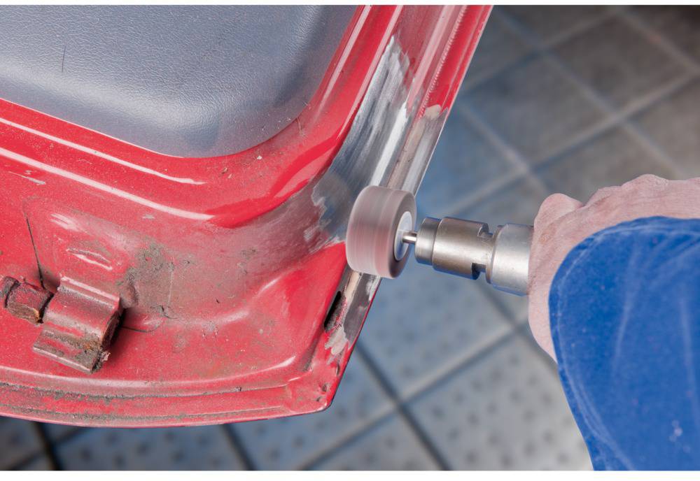 Une personne utilise une perceuse pour enlever la peinture d'une pièce rouge de voiture. Le travail se déroule sur un sol carrelé.