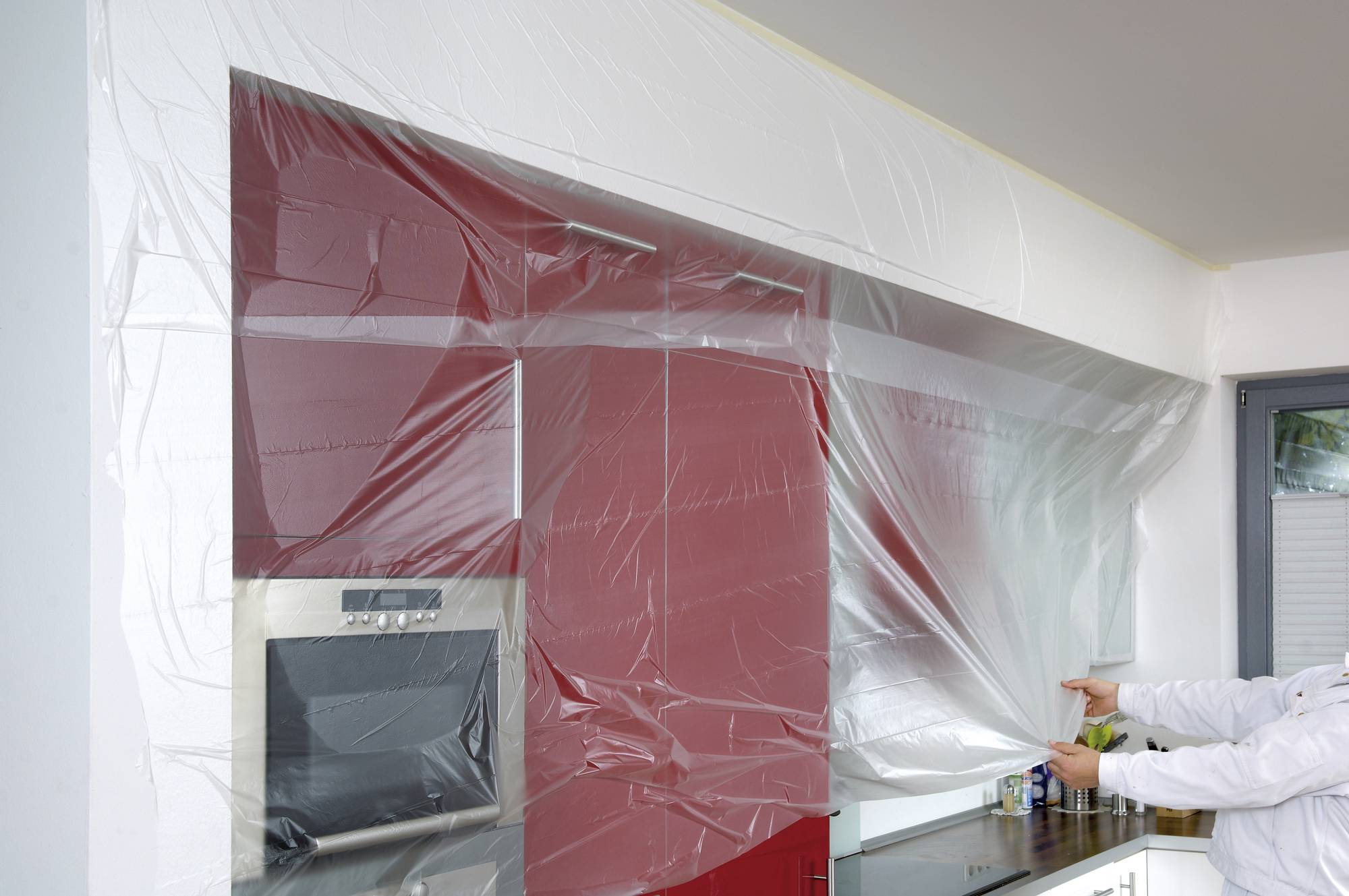 Une personne fixe un film de protection transparent sur un mur de cuisine rouge afin de le protéger de la peinture ou de la poussière.