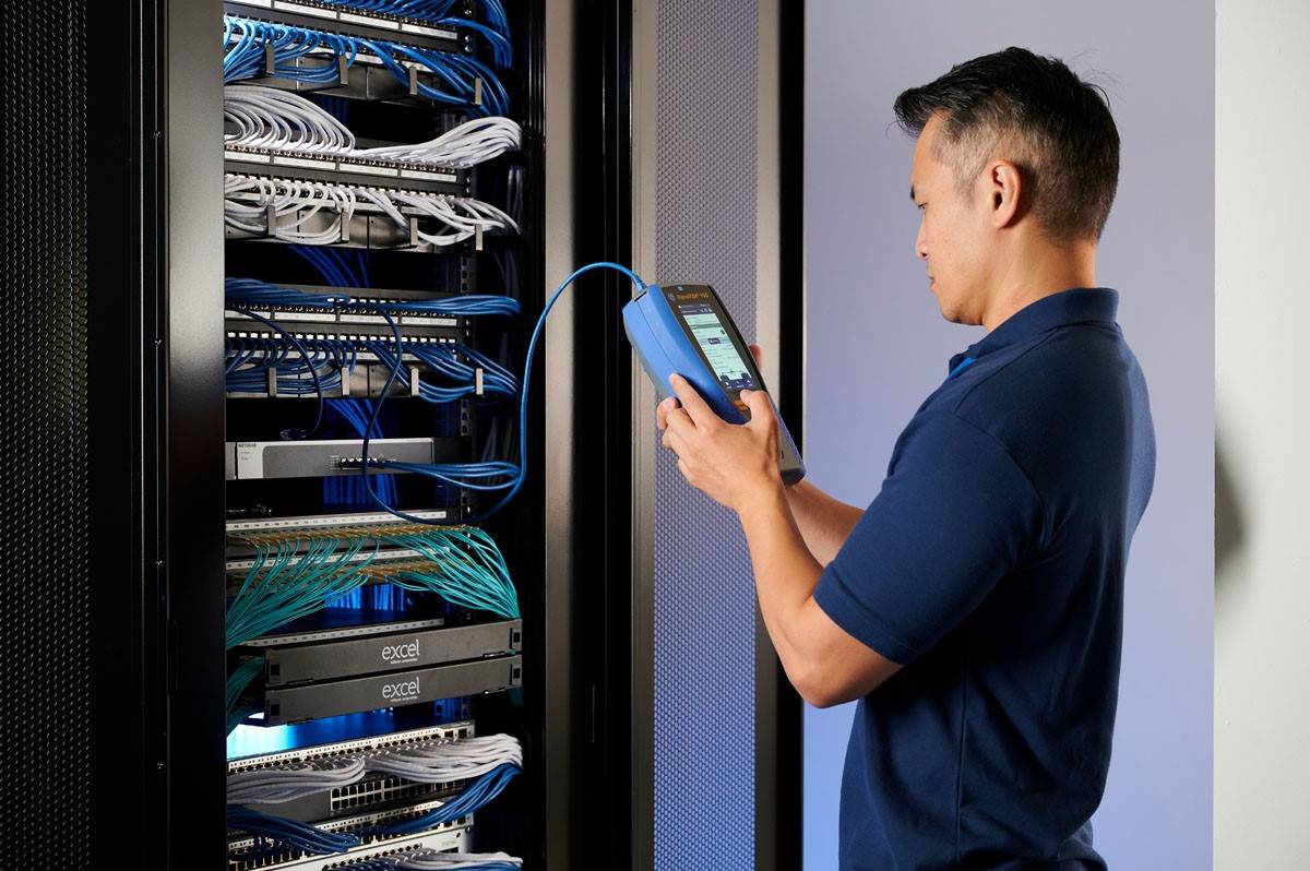 Une personne portant une chemise bleue vérifie les câblages réseau dans une salle de serveurs à l'aide d'un appareil portable.