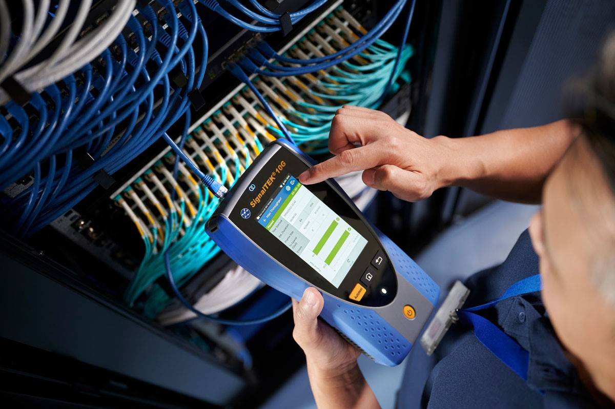 Une personne utilise un appareil de test réseau sur un rack de serveurs pour vérifier la connexion réseau. Les câbles sont soigneusement organisés dans le rack.