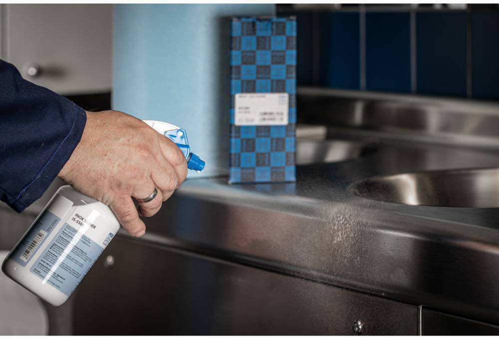 Une main vaporise un produit de nettoyage sur une surface en acier inoxydable dans une cuisine. À l'arrière-plan, une boîte contenant d'autres ustensiles de nettoyage est visible.