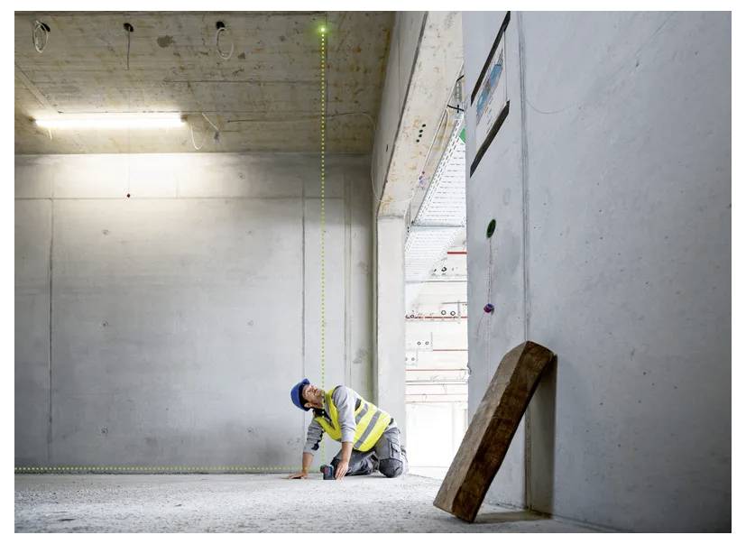 Un ouvrier du bâtiment portant un casque de chantier et un gilet réfléchissant utilise un niveau laser à l'intérieur d'une structure en béton en cours de construction.