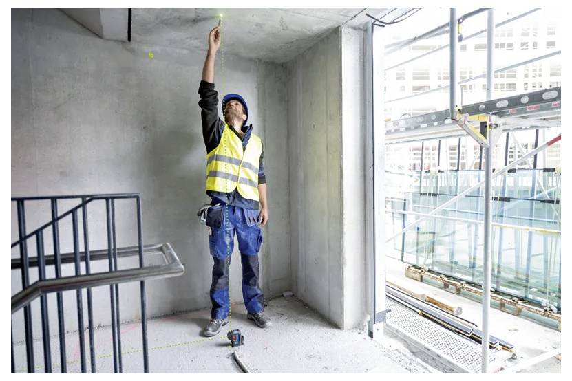 « Un ouvrier du bâtiment portant un gilet de sécurité et un casque marque un point sur le plafond à l'intérieur d'un bâtiment en cours de construction, avec des outils au sol. »