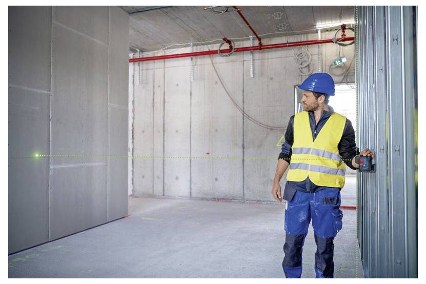 Un ouvrier du bâtiment portant un gilet de sécurité et un casque utilise un niveau laser pour mesurer dans une pièce en béton partiellement construite avec des tuyaux apparents.