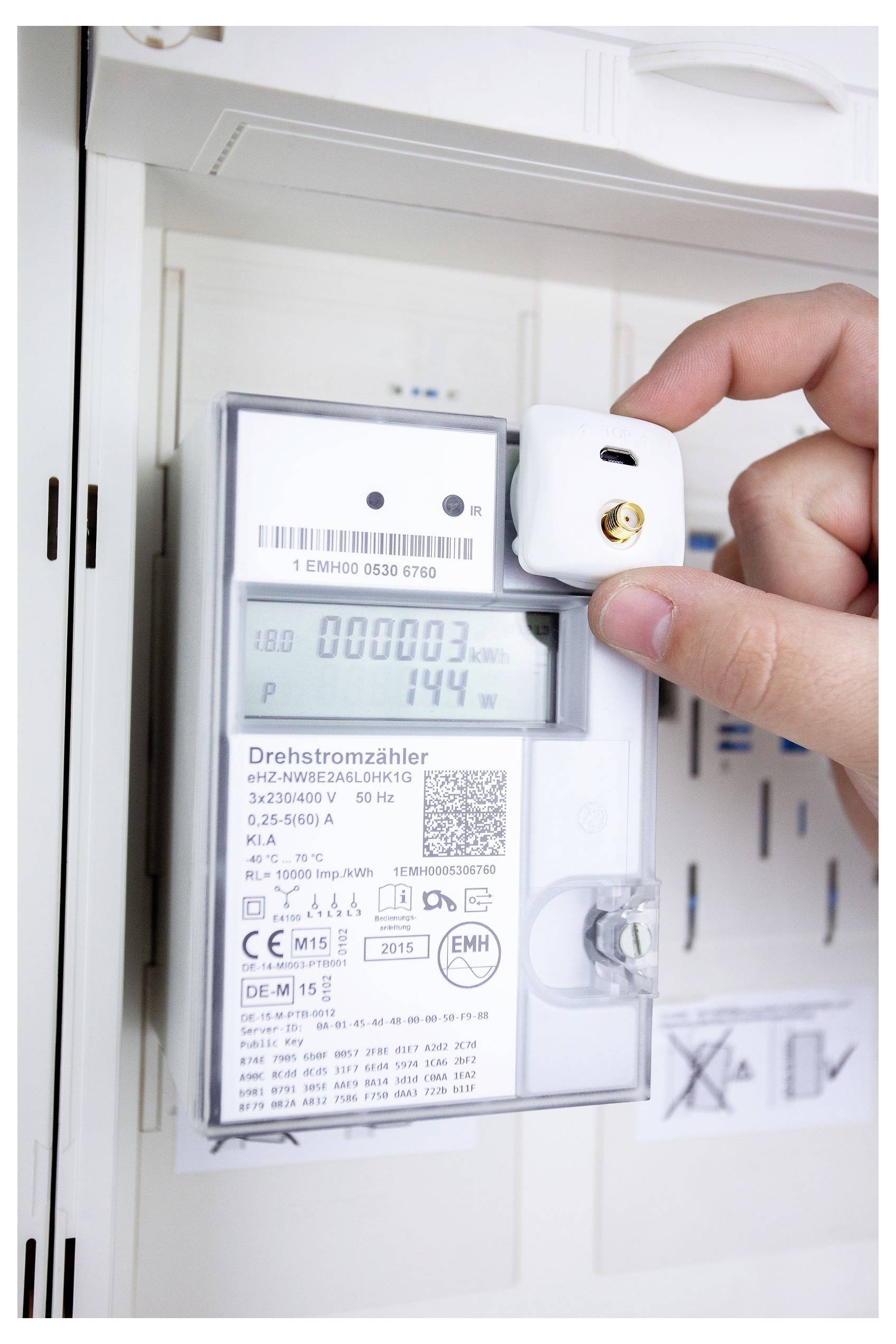 Une main plaçant un petit capteur sur un compteur électrique affichant « 000003 » kWh et « 144 » watts, indiquant la surveillance de la consommation d'énergie.