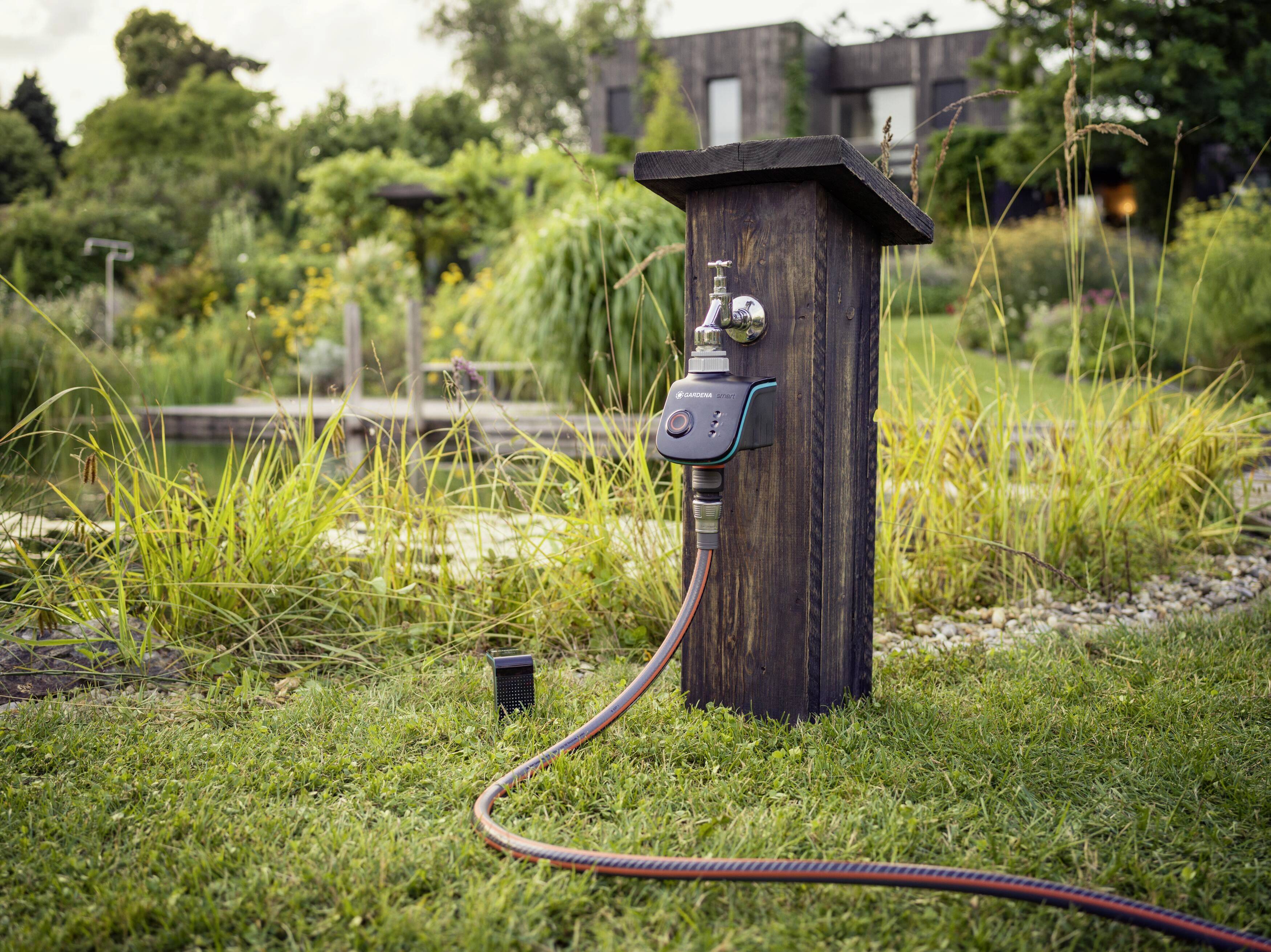 Un tuyau d'arrosage est connecté à un robinet en bois dans un jardin verdoyant. Des arbres et une maison sont visibles en arrière-plan.