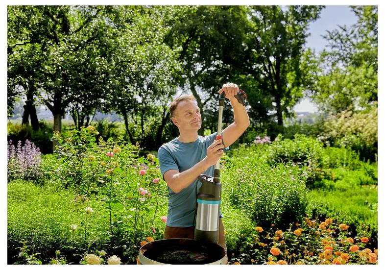 Homme en plein air dans un jardin ensoleillé ajustant un appareil ou une pompe en acier inoxydable à côté d'un parterre de fleurs.