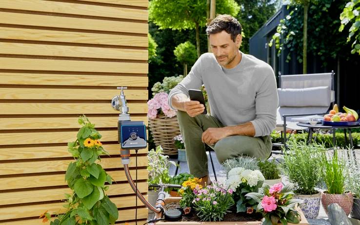 Eine Person in einer Gartenumgebung benutzt ein Smartphone in der Nähe von blühenden Blumen und einem Gartenschlauch, der an einer Holzwand befestigt ist. Im Hintergrund befindet sich eine gemütliche Sitzgelegenheit.