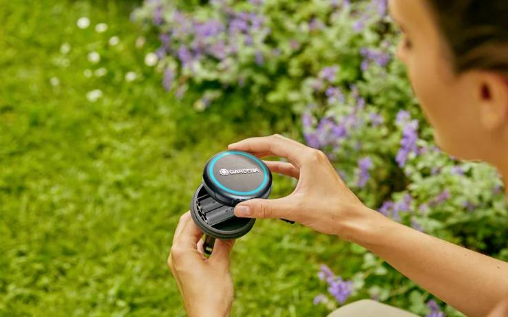 Une personne tient un appareil rond étiqueté 'GARDENA' près d'un jardin avec de la lavande en fleurs, suggérant un usage pour l'entretien ou la maintenance du jardin.