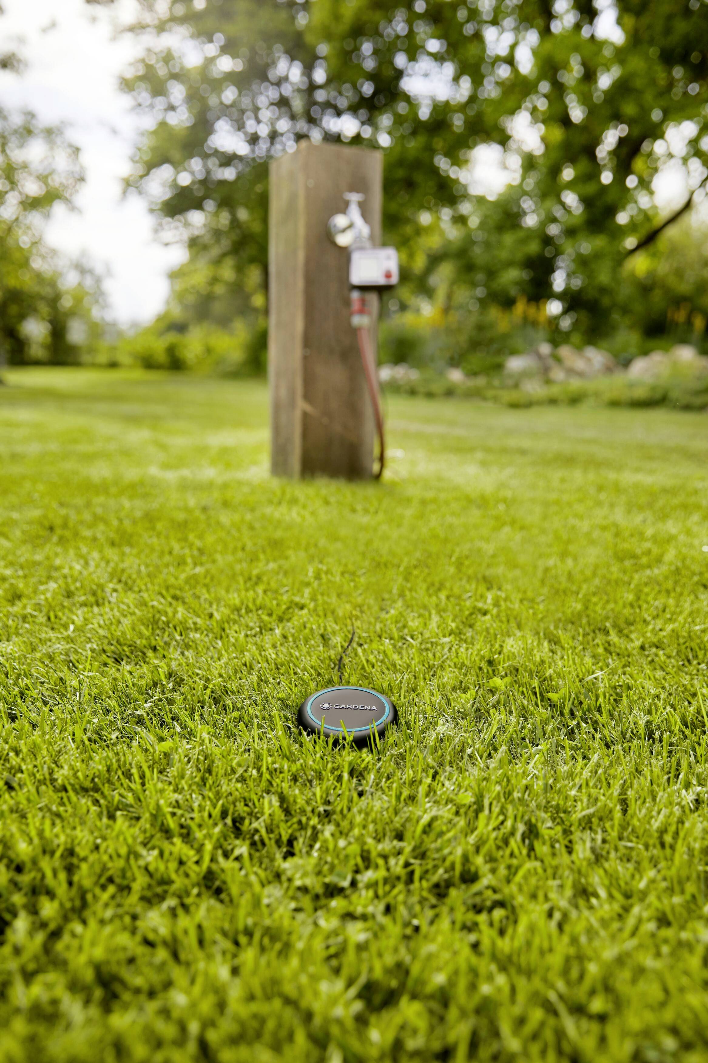 Un capteur est posé dans l'herbe devant un système d'irrigation dans un pré. À l'arrière-plan, on peut voir un poteau en bois avec un robinet.
