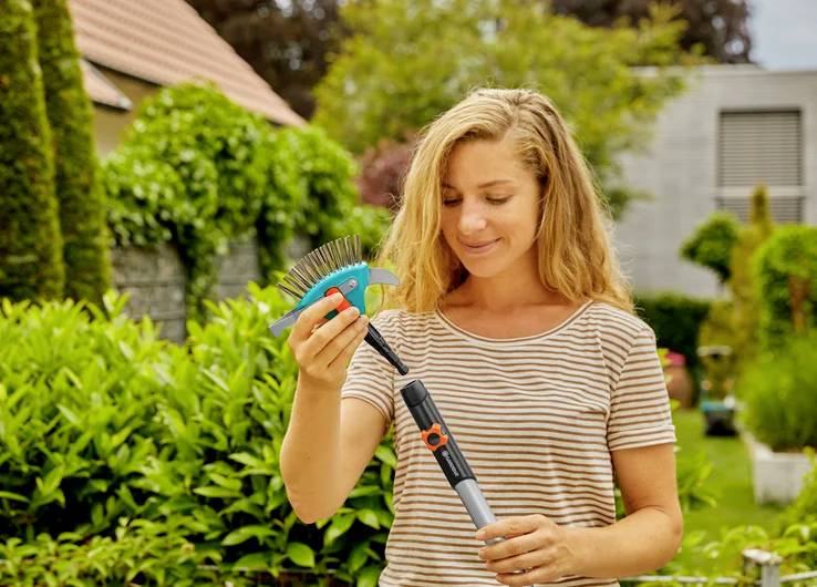 Une femme dans un jardin tenant un petit râteau, entourée de plantes vertes luxuriantes, suggérant des activités de jardinage.
