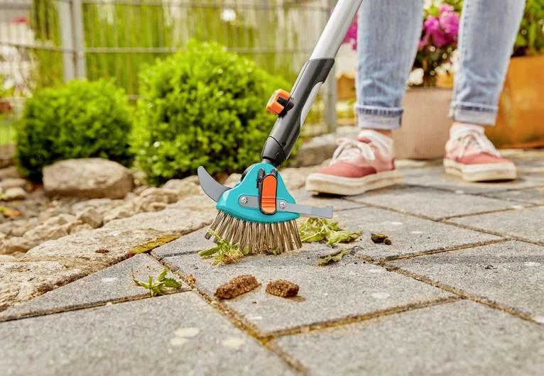 Une personne utilise un outil de jardin pour nettoyer la mousse et les débris d'un chemin de pierre. Des fleurs roses et des buissons verts sont visibles en arrière-plan.