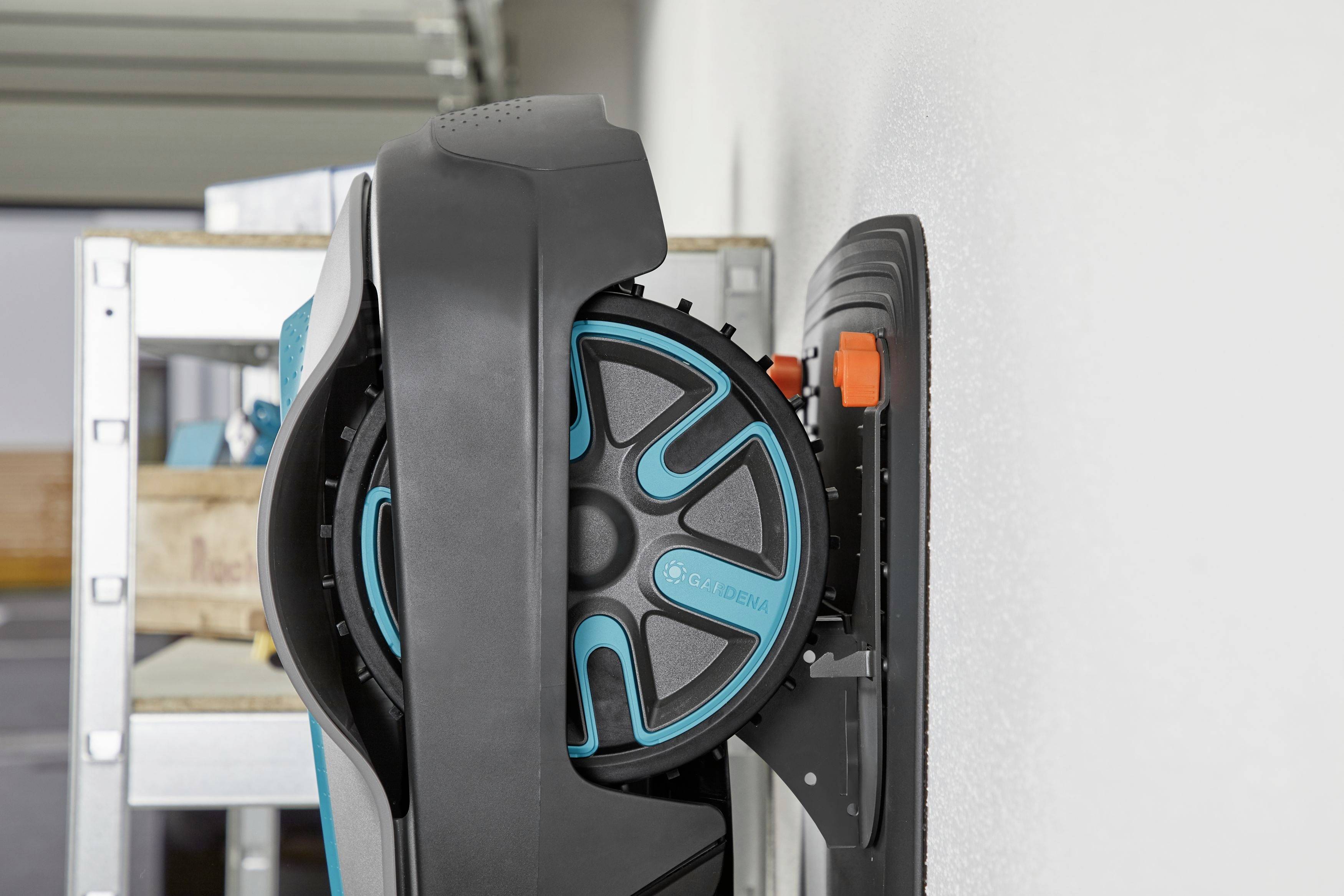 Un robot tondeuse est fixé à un support mural dans un garage. Des étagères avec des outils sont visibles en arrière-plan.