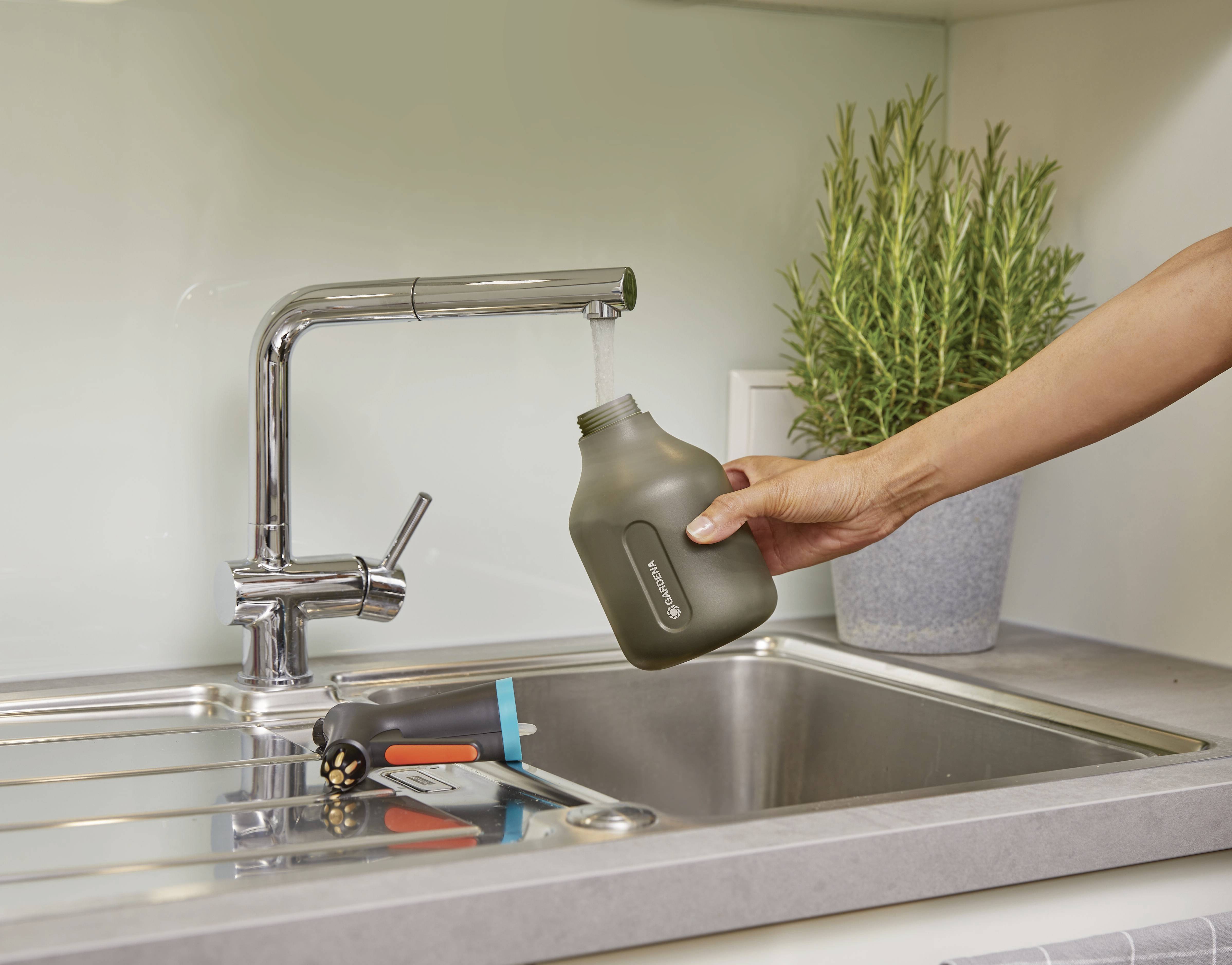 Une main tient une bouteille d'eau grise sous un robinet qui coule dans une cuisine. À côté se trouvent un arrosoir noir et une plante verte.