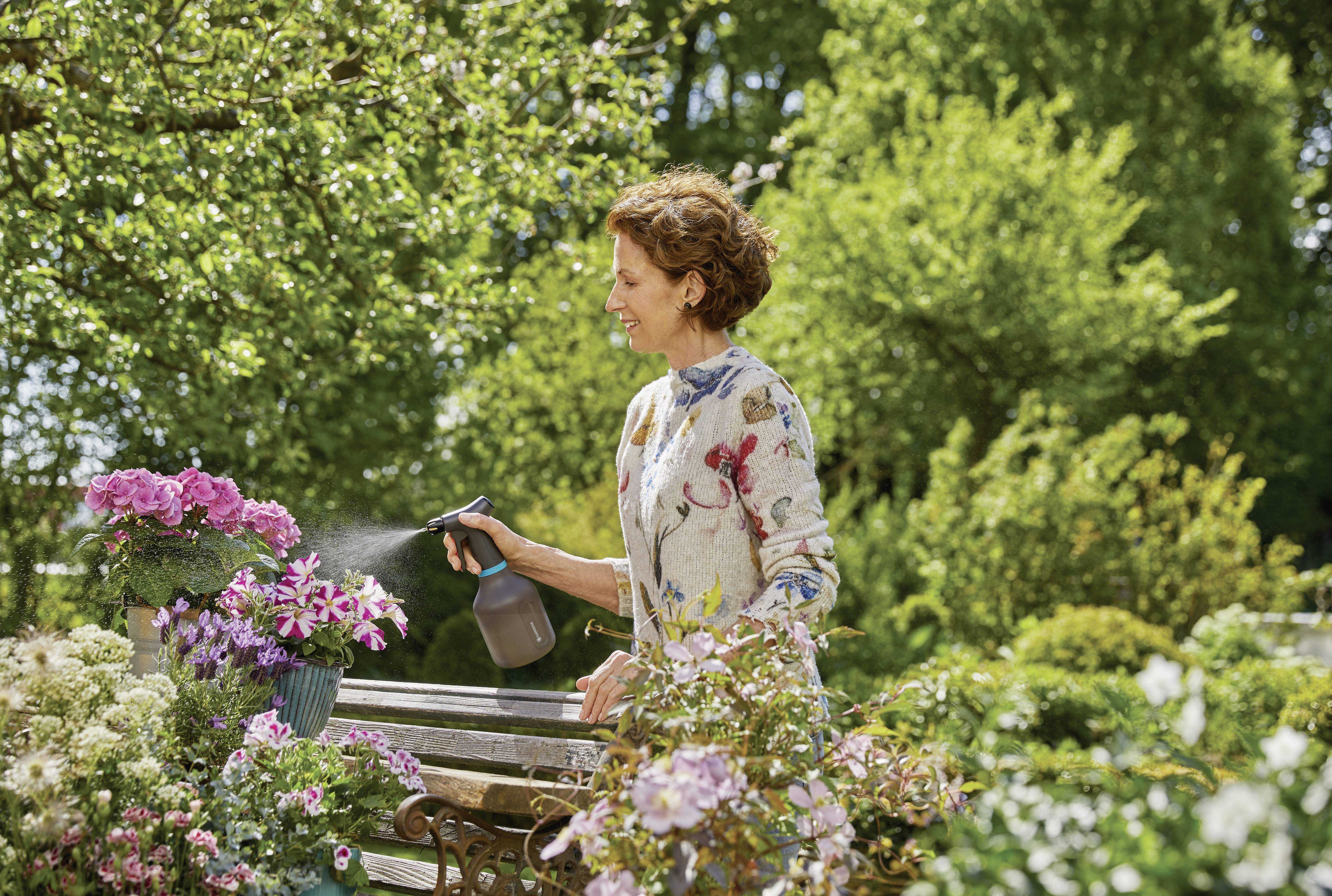 Une femme dans un jardin arrose des fleurs avec de l'eau. Elle porte un chemisier à motifs floraux. Autour d'elle, il y a des arbres verts et des plantes en fleurs.