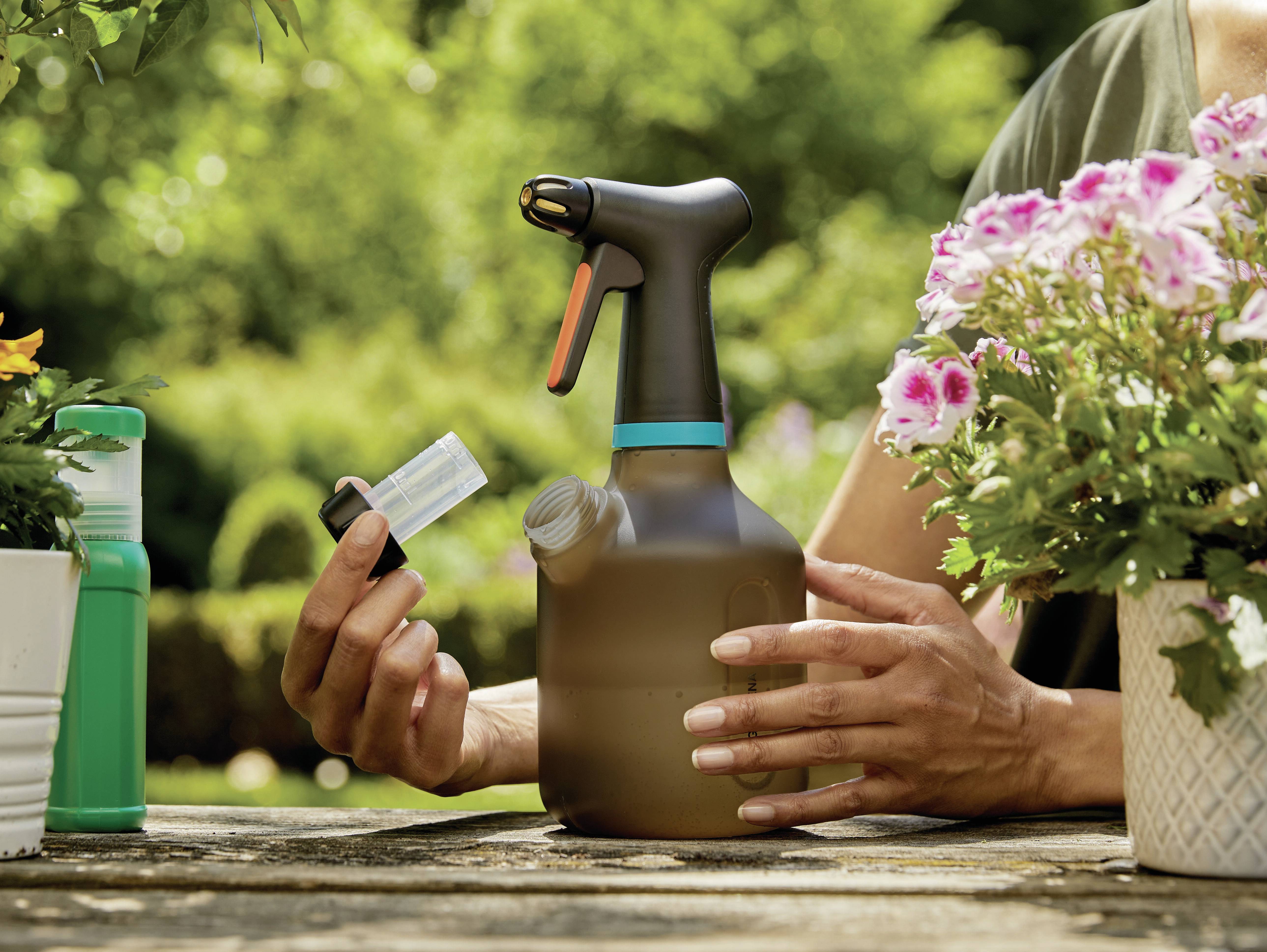 Une personne tient un pulvérisateur et une petite bouteille dans un jardin rempli de plantes en fleurs.
