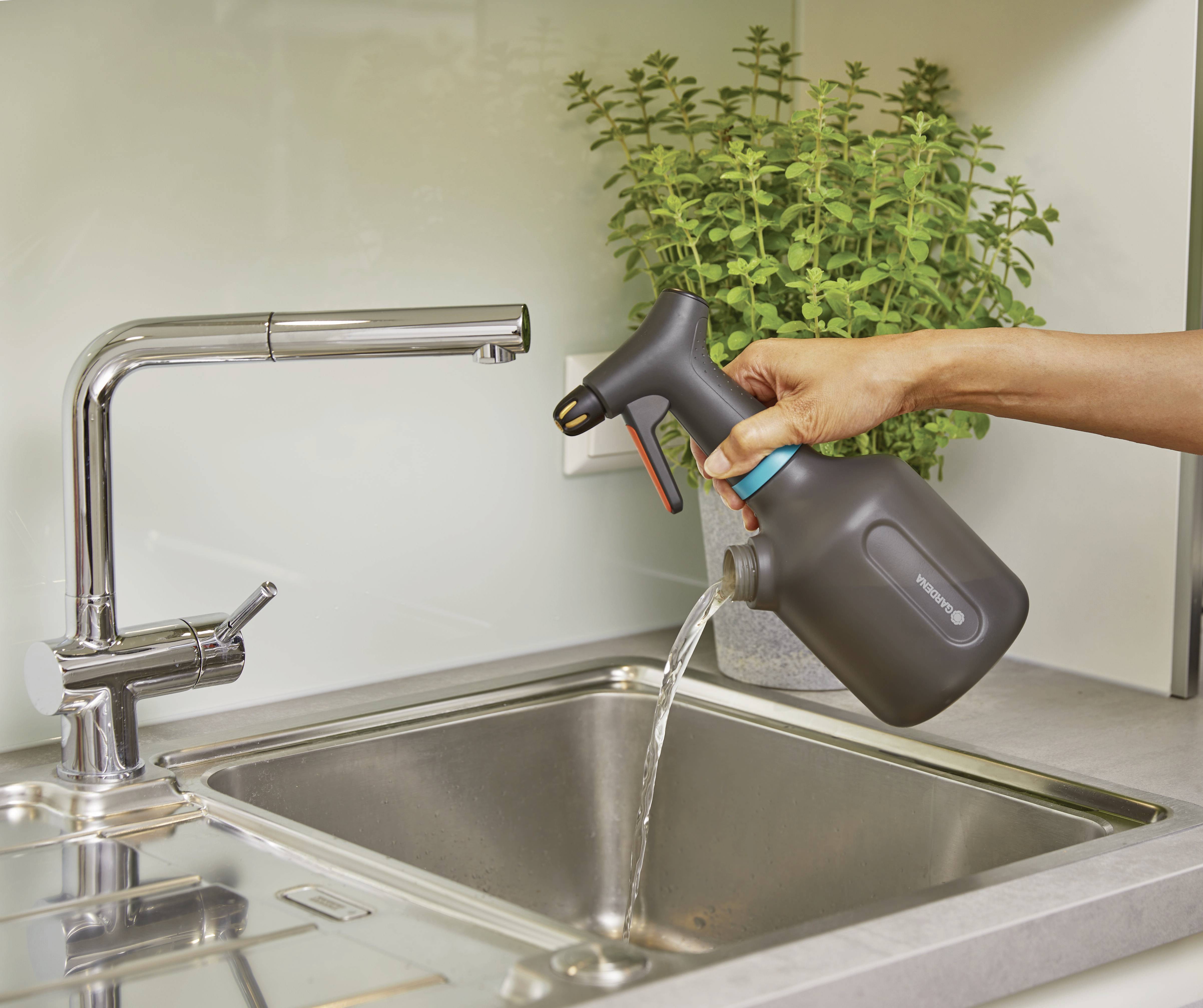Une main verse de l'eau d'un arrosoir dans un lavabo. Une plante est visible en arrière-plan.