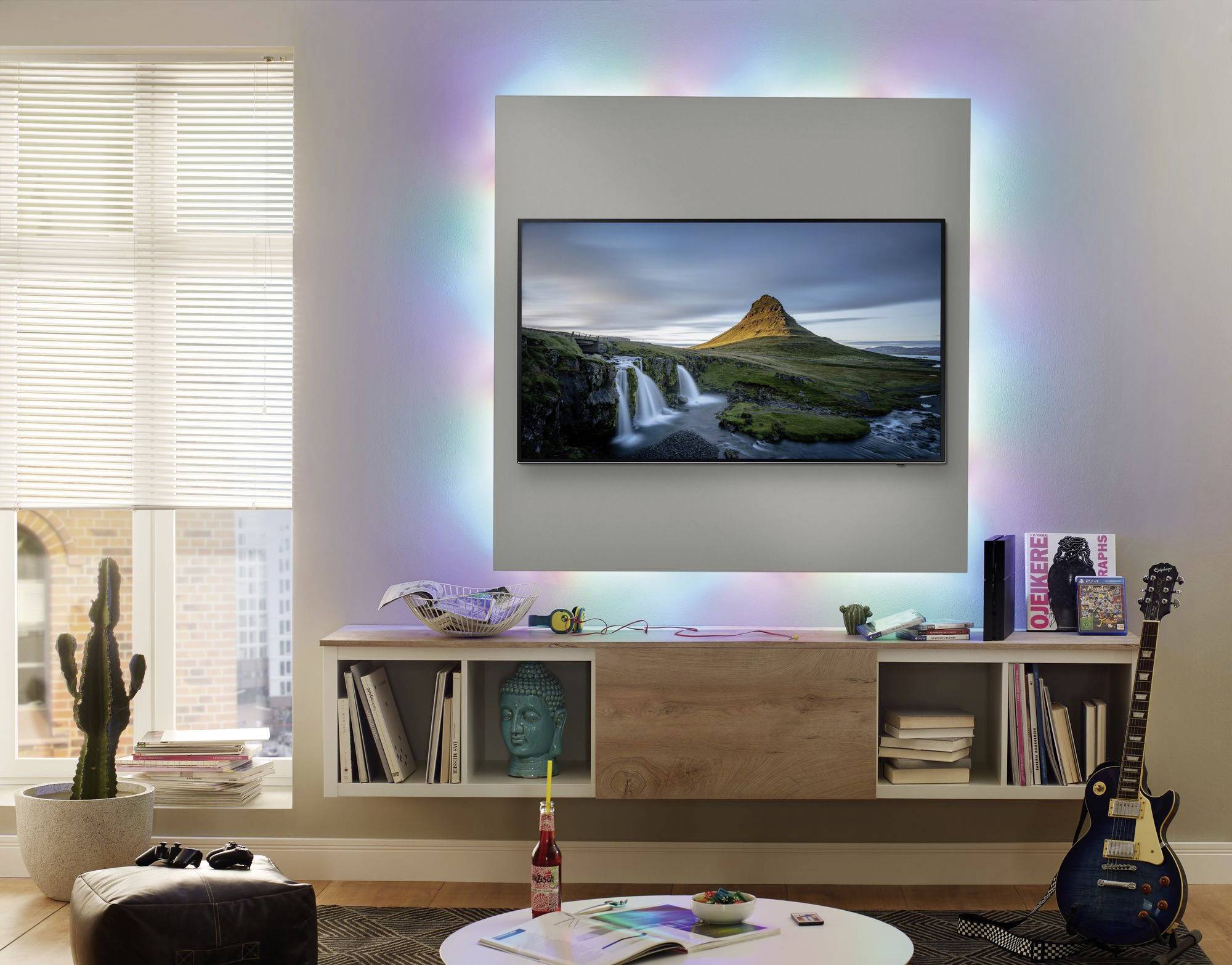 Un salon avec un grand téléviseur au mur montrant un paysage. Un cactus et une guitare sont placés à gauche et à droite.