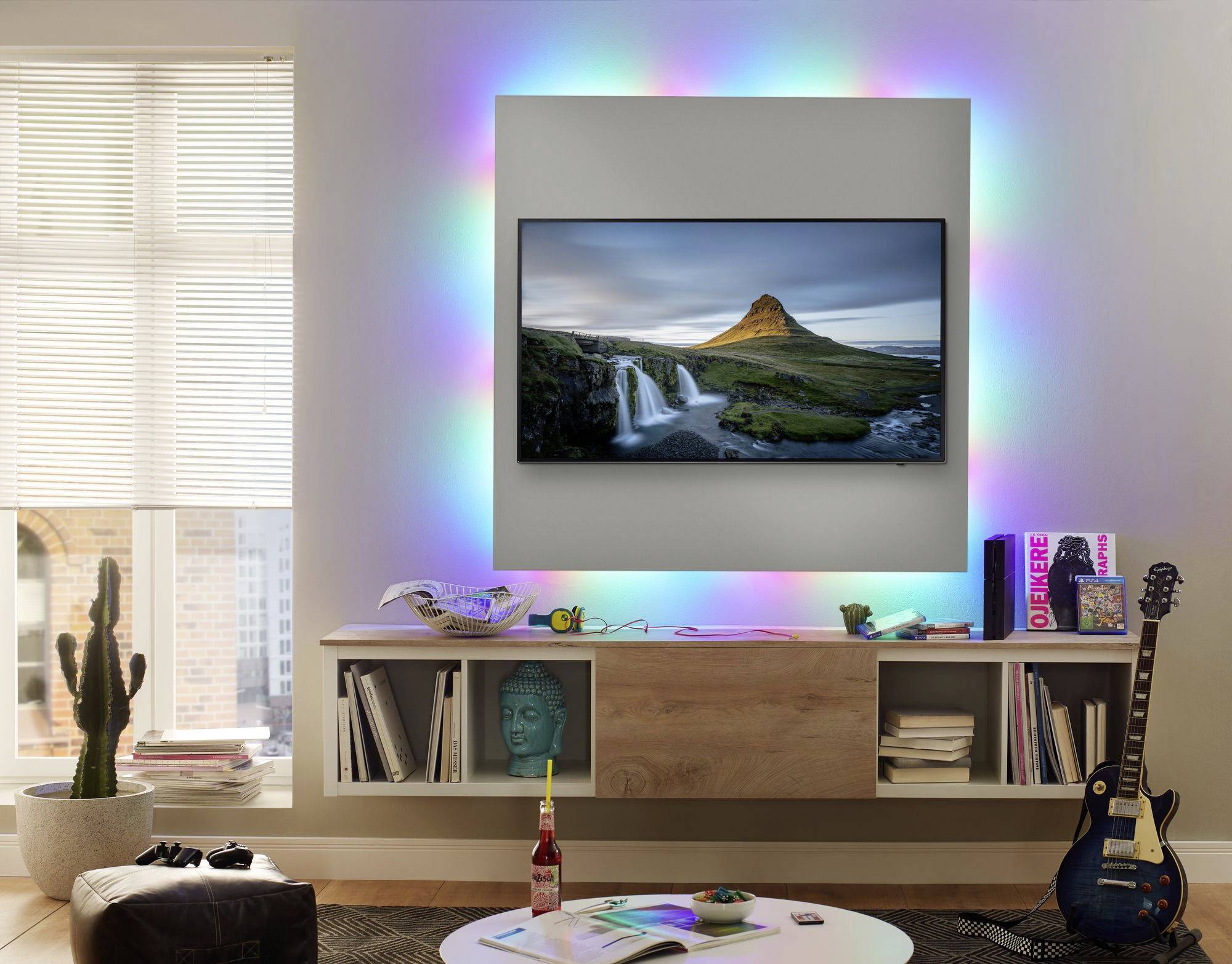 Écran plat accroché au mur avec une image de paysage, entouré de lumières LED colorées. En dessous, des livres, une guitare et des décorations sont disposés.