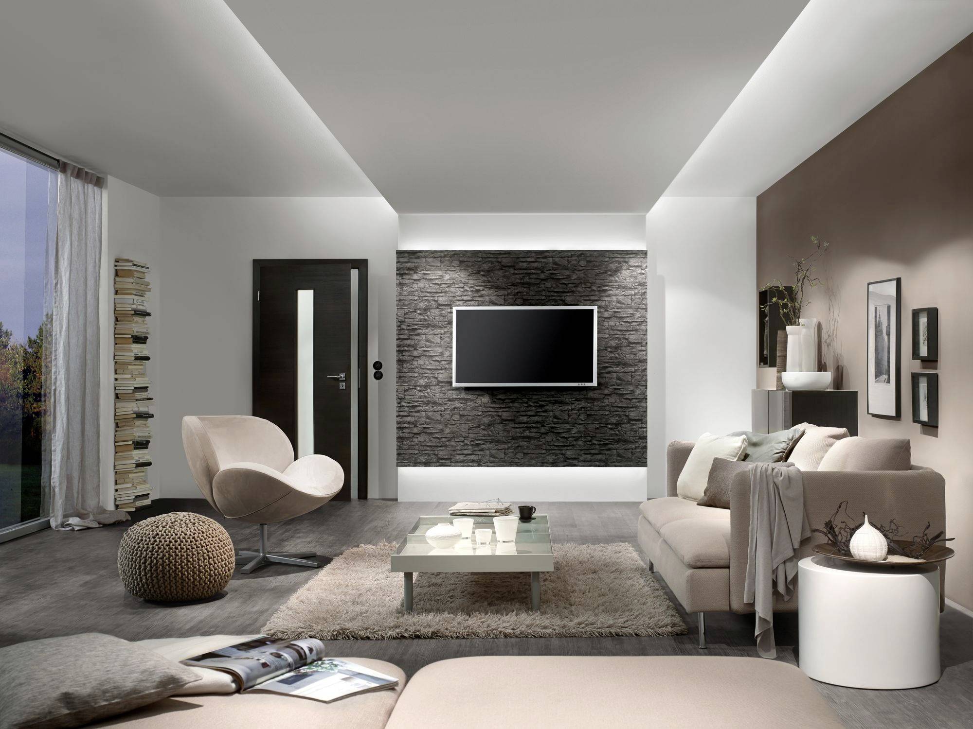 Salon moderne avec des canapés beiges, un téléviseur à écran plat contre un mur en pierre et une grande fenêtre sur la gauche.