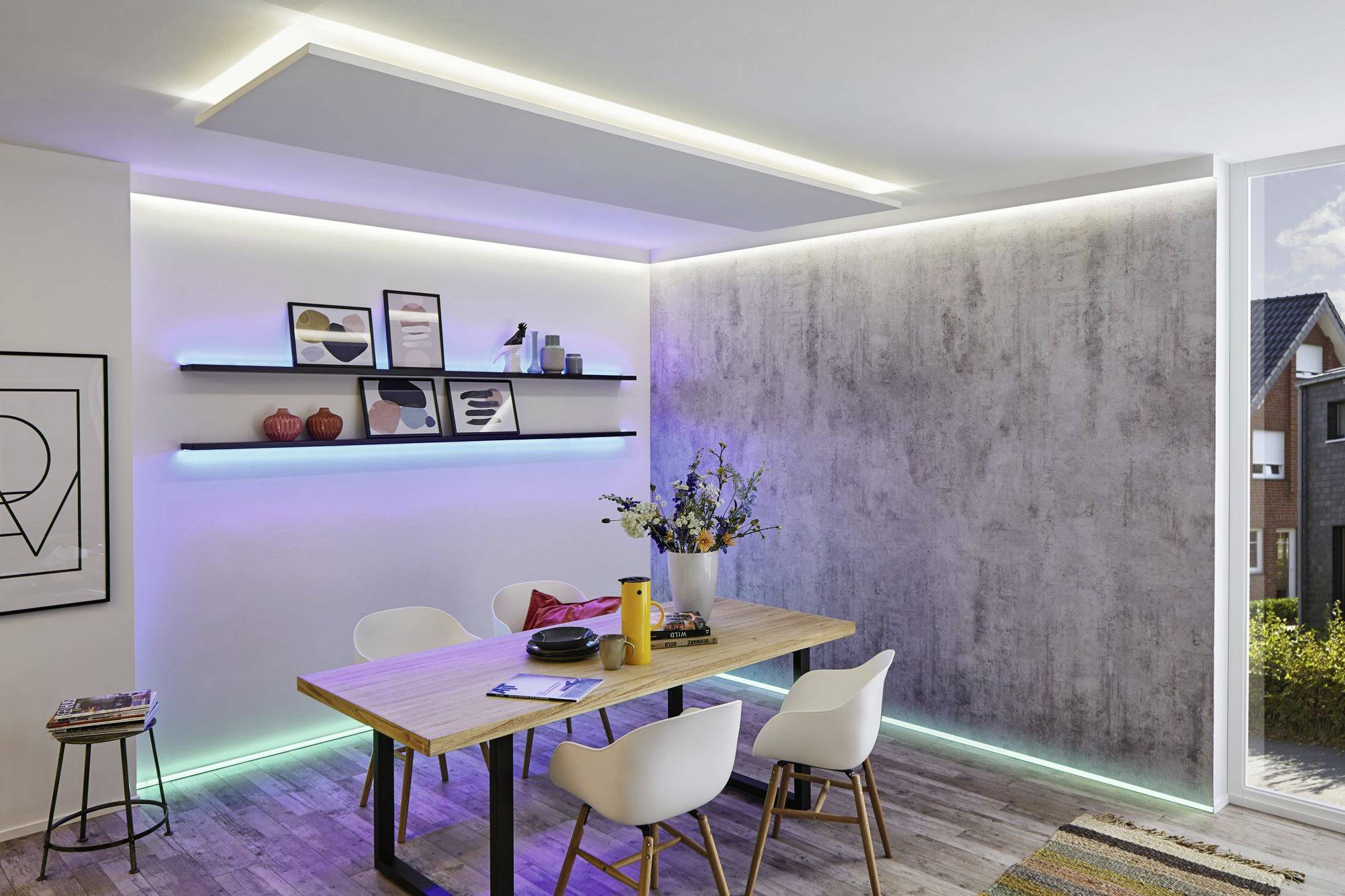 Salle à manger avec une table moderne et des chaises blanches, éclairée par des bandes LED. Des étagères avec des objets décoratifs et des fleurs sur la table.