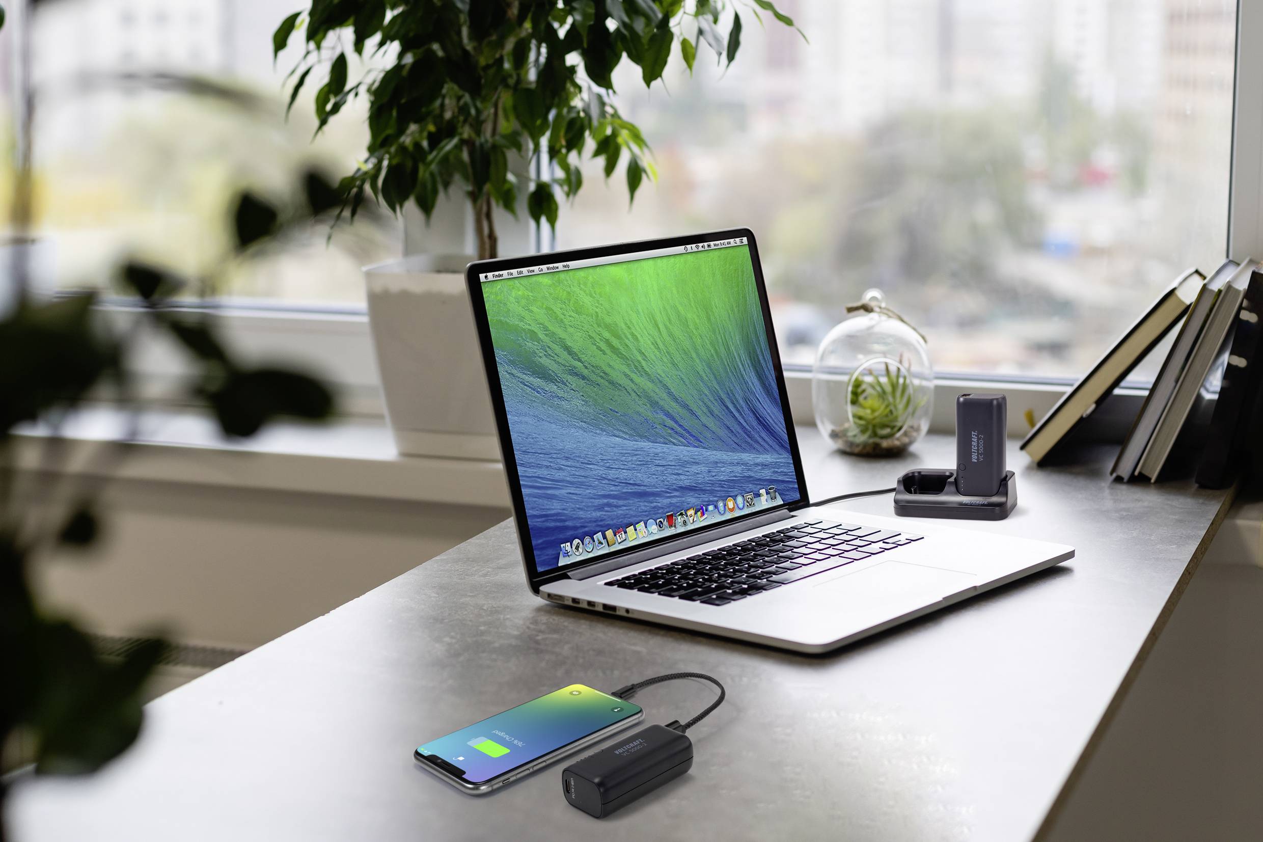 Un ordinateur portable posé sur une table dans un bureau moderne avec vue sur la ville. À côté, un smartphone et une batterie externe.