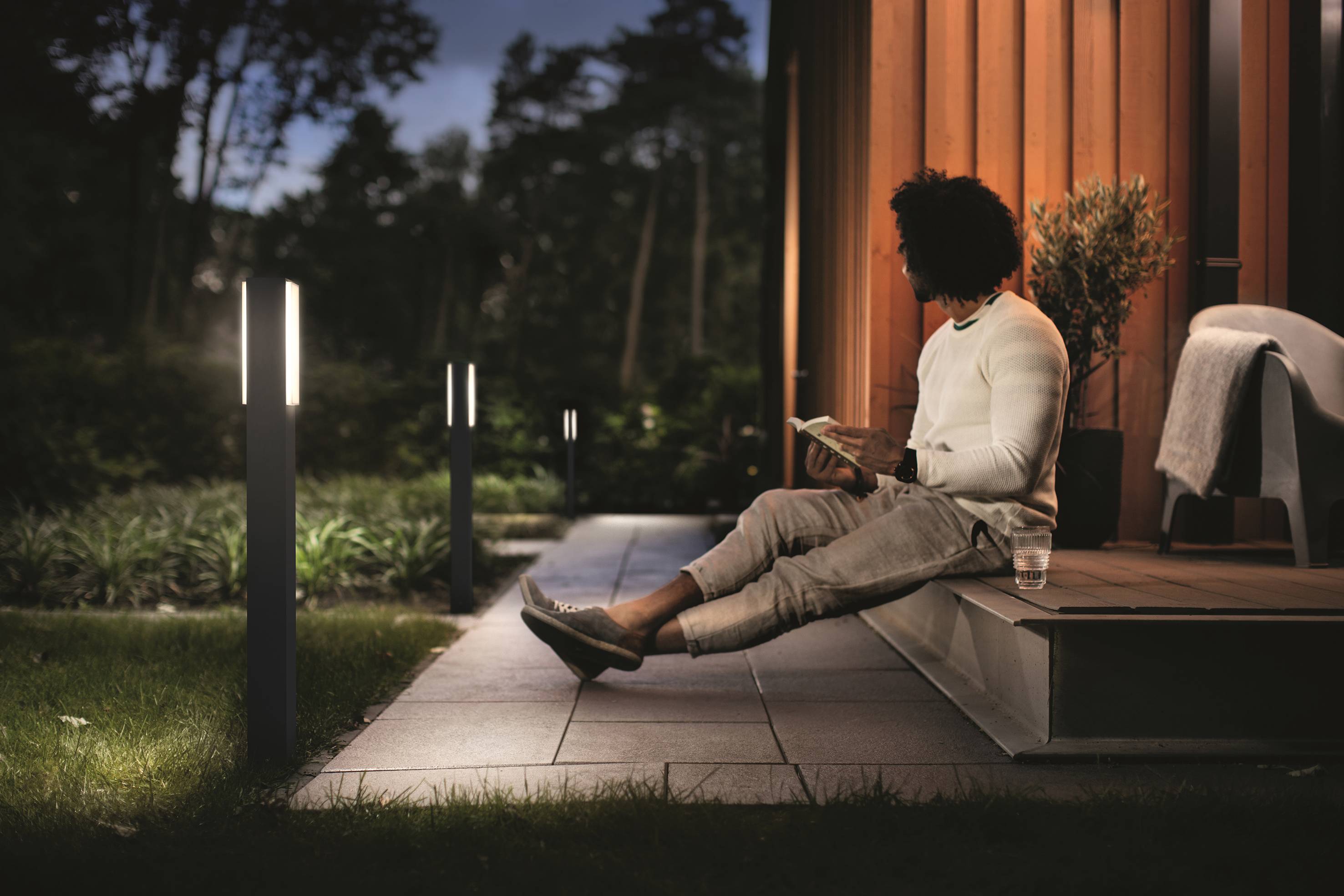 Un homme est assis sur une terrasse et lit au crépuscule. Un éclairage de jardin moderne illumine le chemin et les arbres environnants sont visibles.