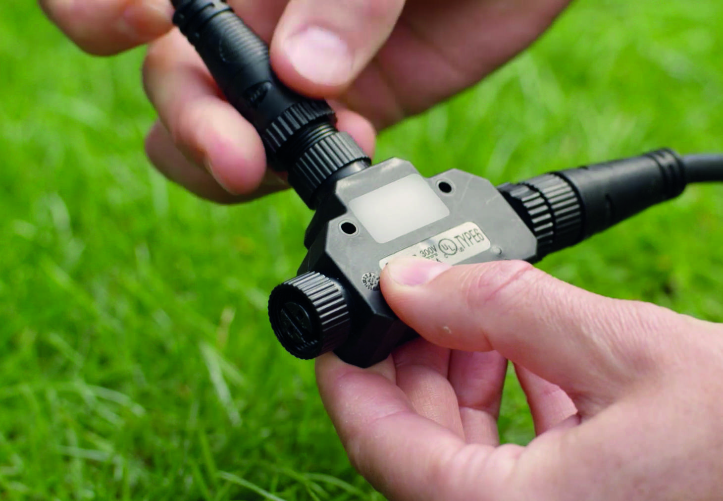 Une personne tient un câble noir avec des connecteurs dans ses mains. L'arrière-plan montre de l'herbe verte.