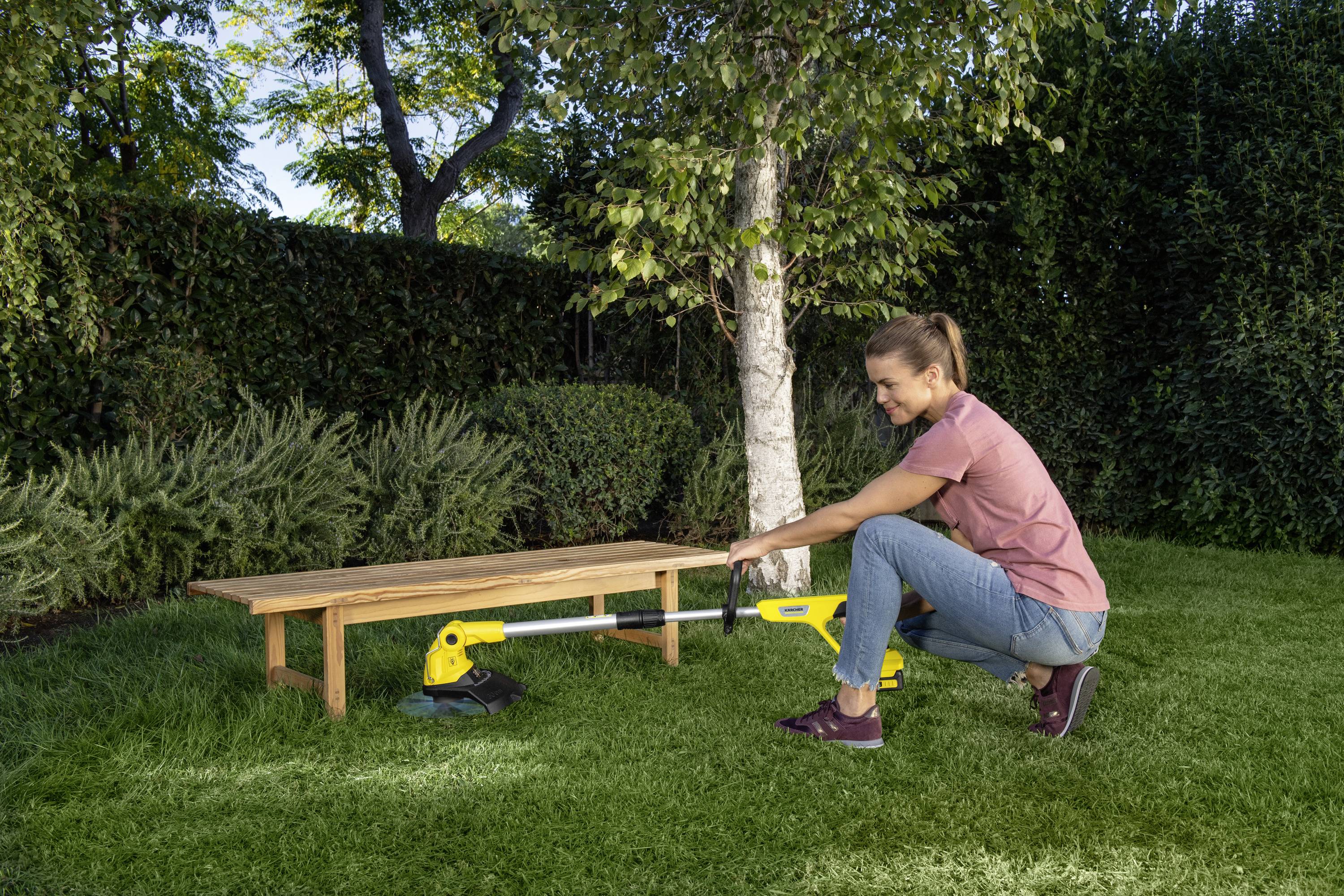 Une personne est agenouillée sur l'herbe dans un jardin et utilise un appareil électrique pour enlever les mauvaises herbes. Des arbres sont visibles en arrière-plan.