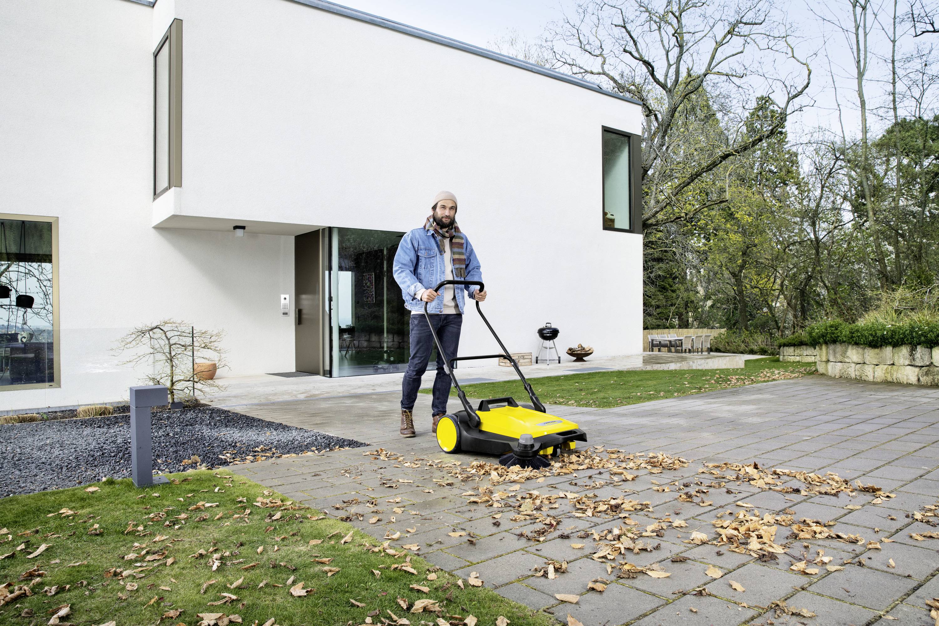 Une personne utilise un balai mécanique jaune pour enlever des feuilles d'un chemin pavé devant une maison moderne.