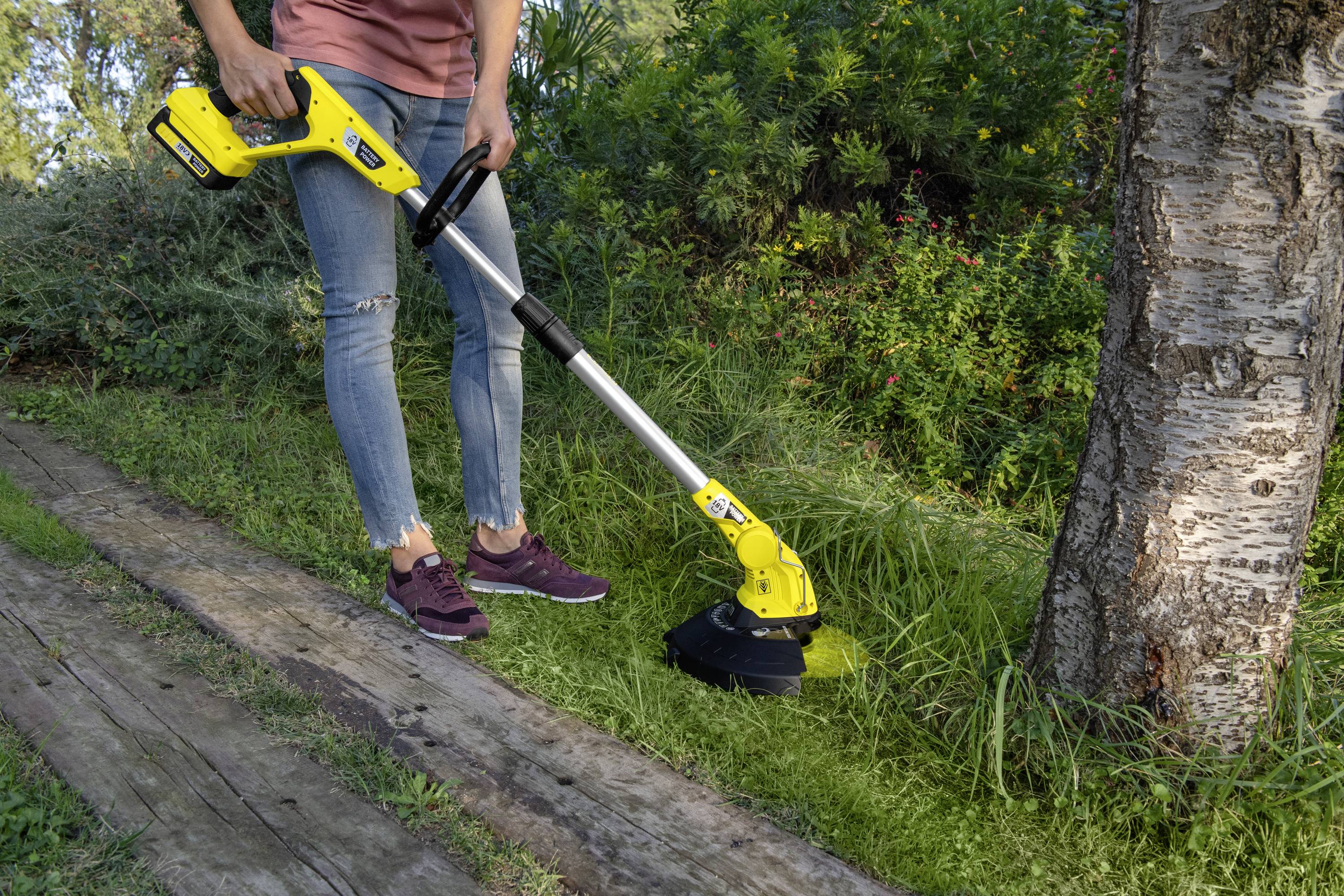 Une personne portant un jean et des baskets utilise un coupe-bordures jaune pour tondre l'herbe au bord d'une terrasse en bois à côté d'un arbre.