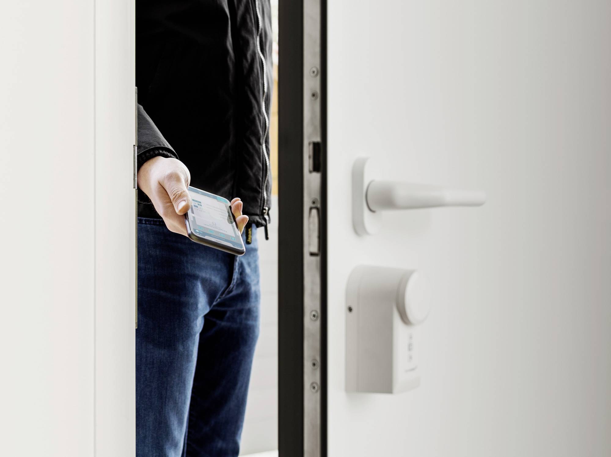 Une personne ouvre une porte en tenant un smartphone à la main. Sur la porte, on peut voir une serrure électronique.