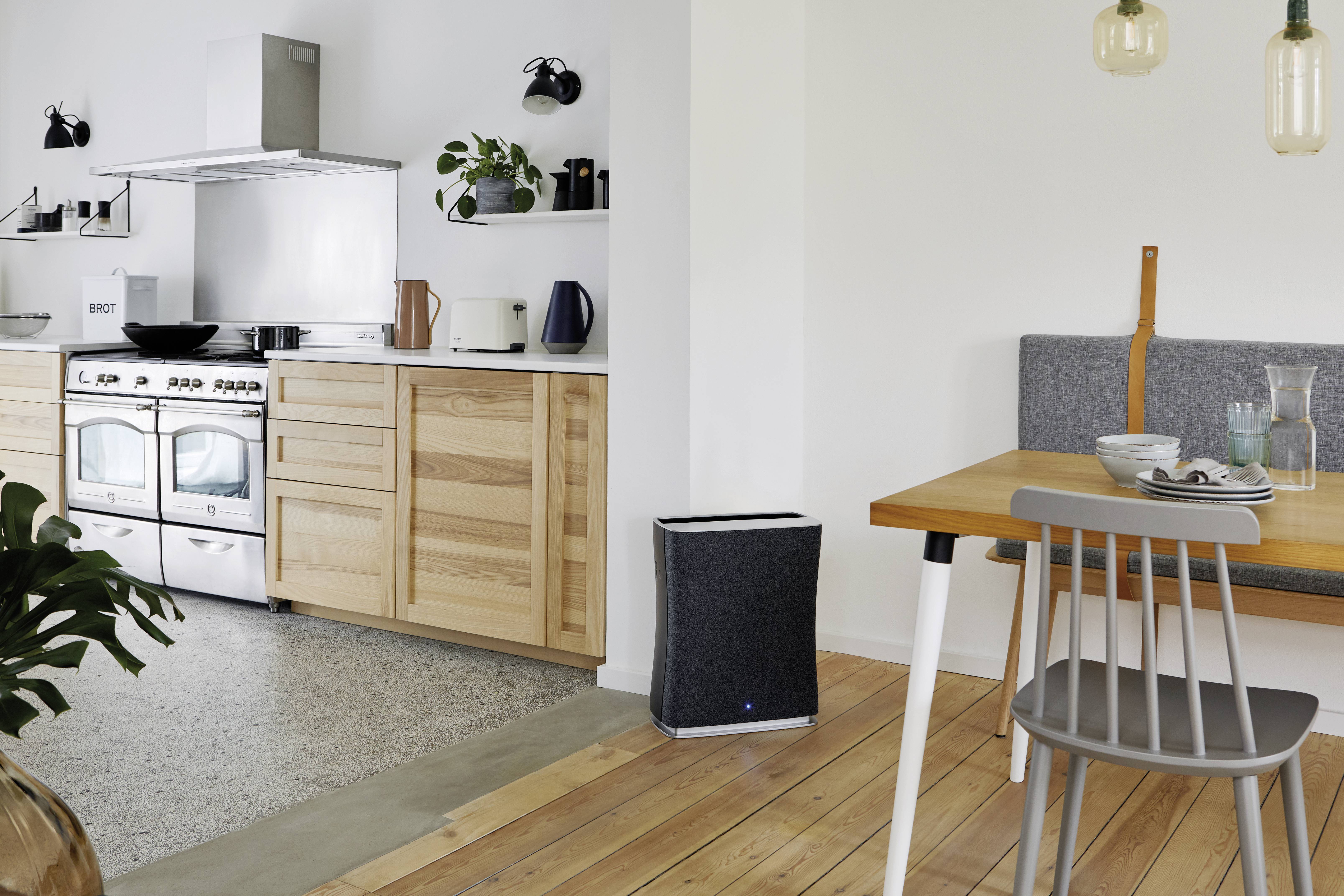 Cuisine moderne avec des murs blancs et un sol en bois. À gauche, une ligne de cuisine avec une cuisinière, à droite une table à manger avec une chaise. Décorations discrètes.