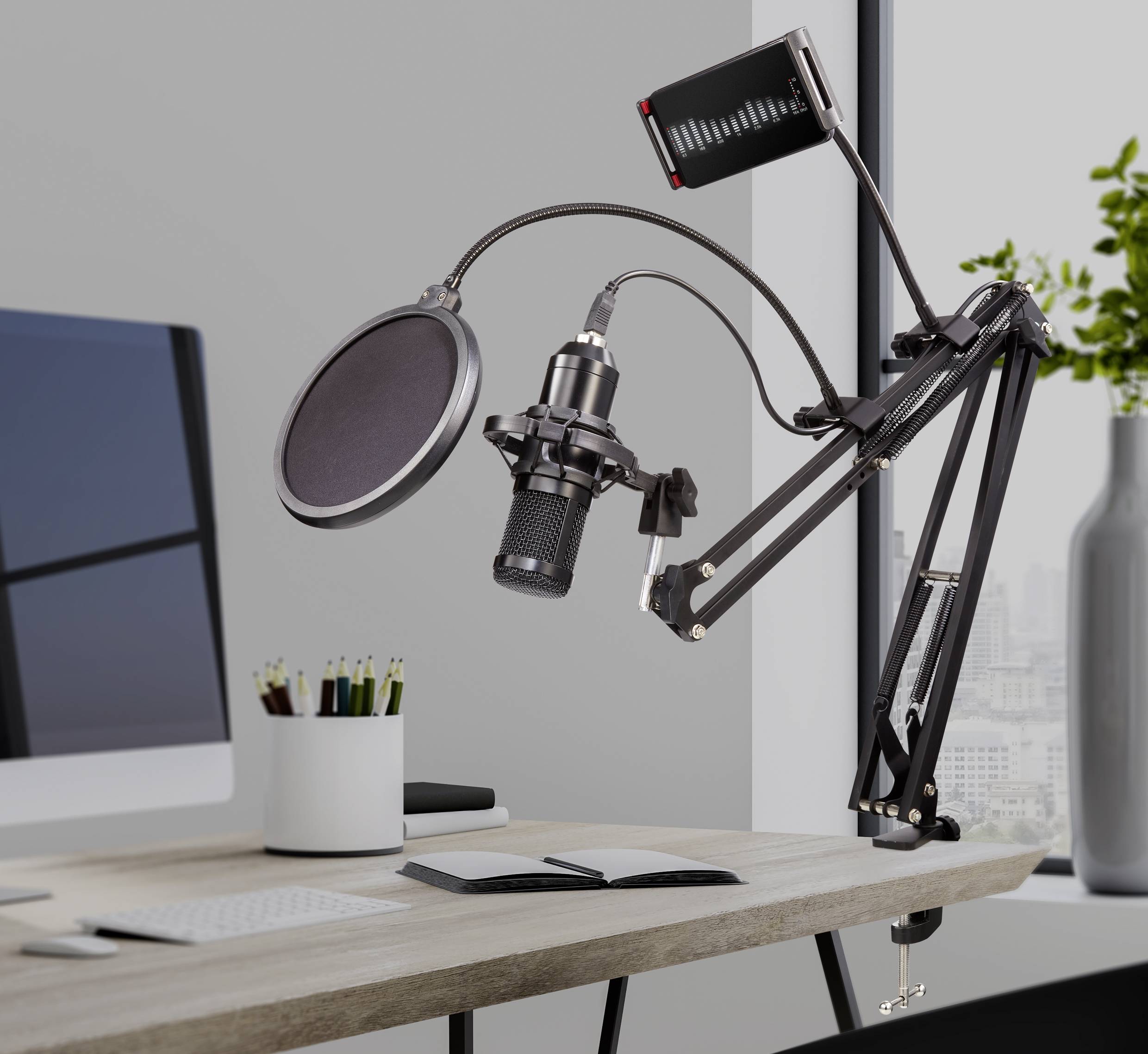 Microphone avec filtre anti-pop sur un bras articulé, à côté d'un écran et de cahiers sur un bureau, pour des enregistrements de podcast.