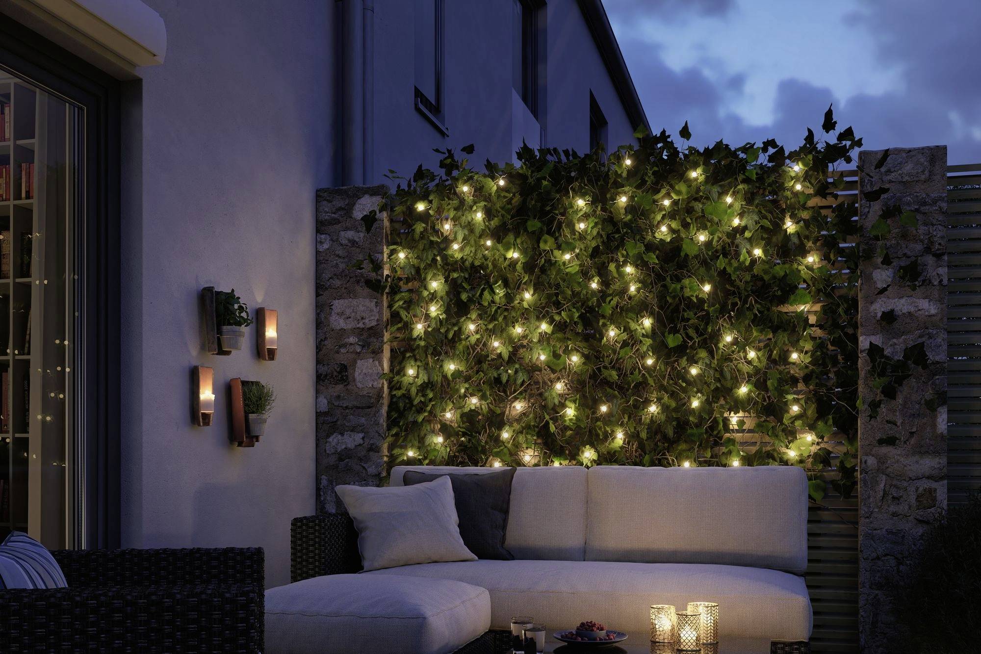 Terrasse accueillante au crépuscule, décorée avec une guirlande lumineuse et des coussins sur un canapé. Des bougies dans des verres complètent l'éclairage chaleureux.