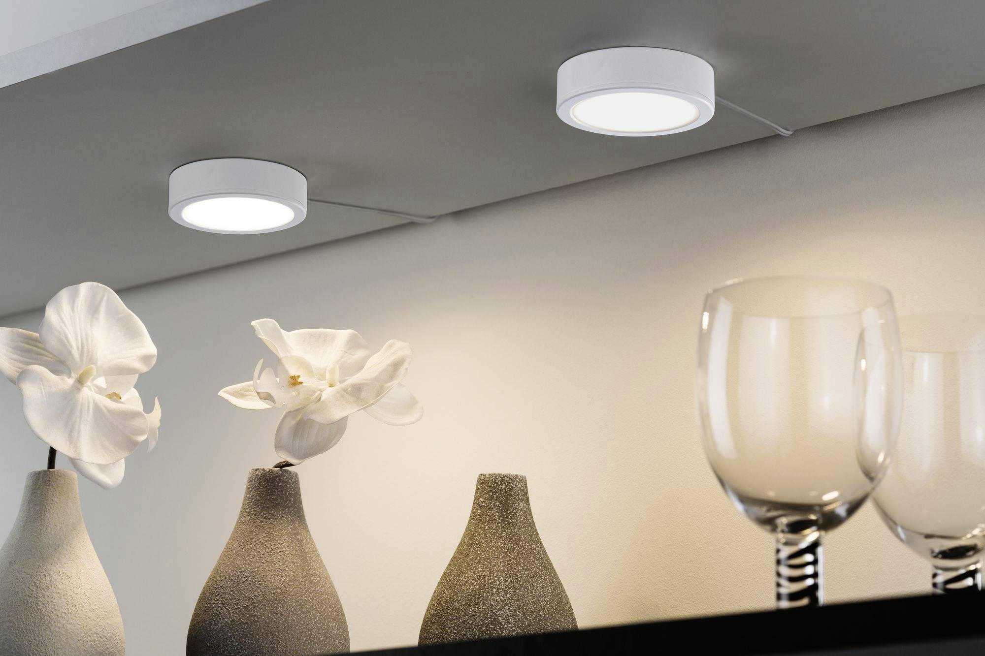 Deux appliques plafonnières blanches et rondes illuminent des vases décoratifs et deux fleurs blanches sur une étagère. Décoration intérieure moderne et élégante.