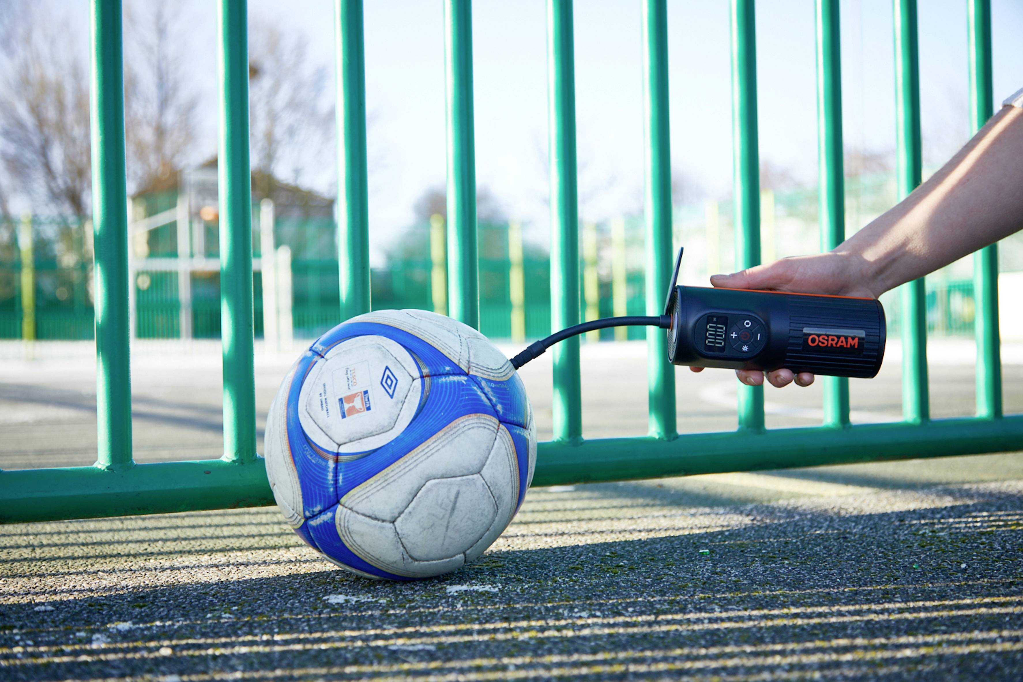 Une main tient une pompe à air Osram fixée à la valve d'un ballon de football. Le ballon est posé sur un terrain de sport entouré d'une clôture verte.