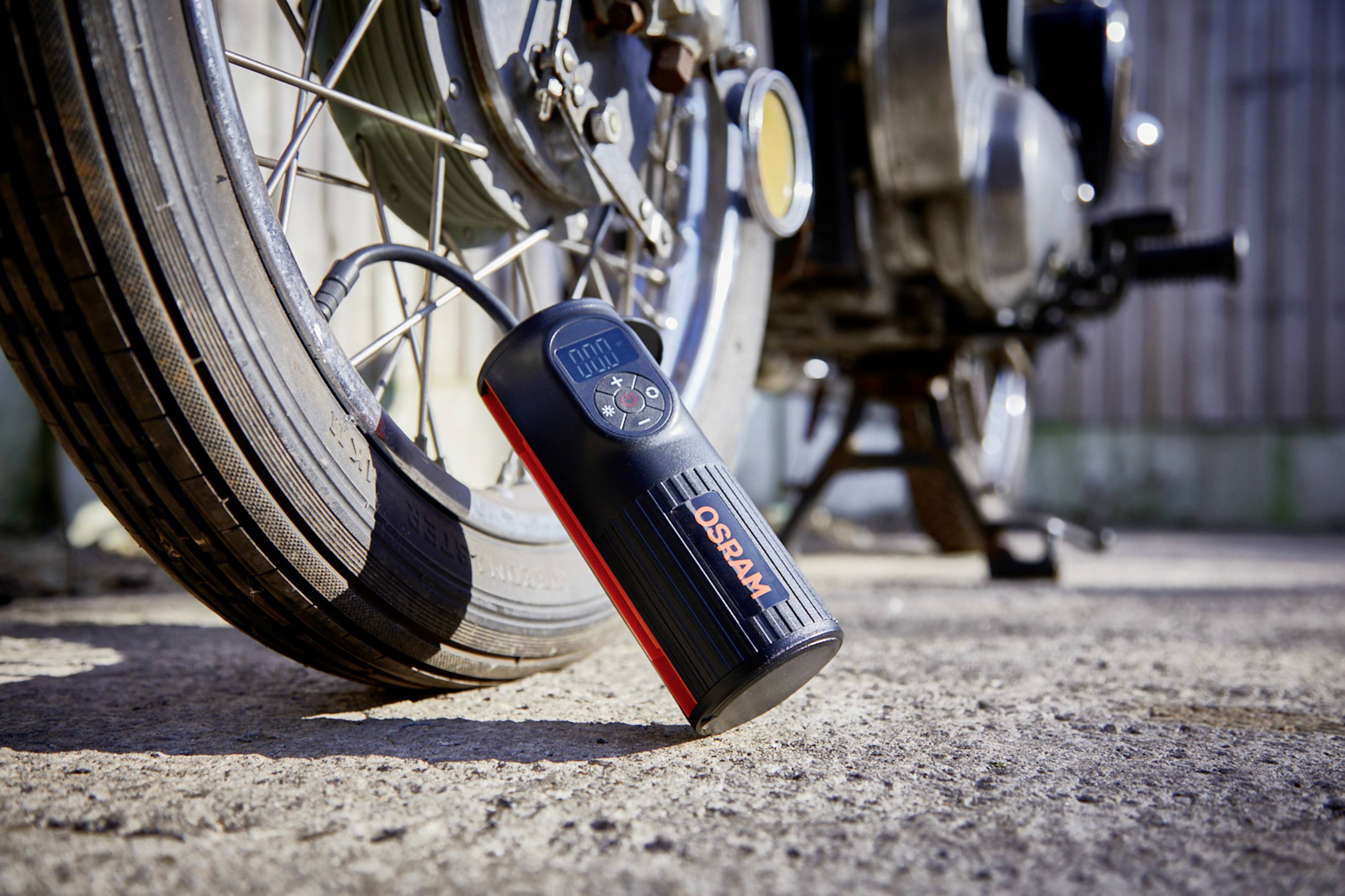 Un compresseur portable de pneus est placé à côté d'un pneu de moto en train de se gonfler. L'écran affiche 0,00 psi. Le soleil brille sur la scène.
