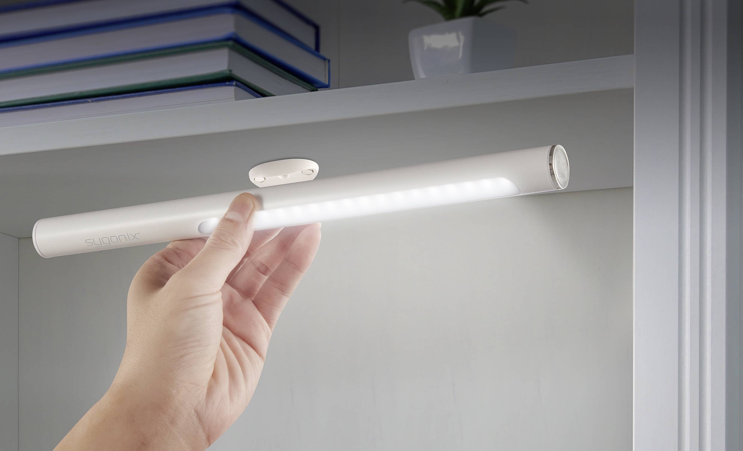 Une main tient une veilleuse LED mince et alimentée par batterie sous une étagère. À l'arrière-plan, on aperçoit des livres et une plante.