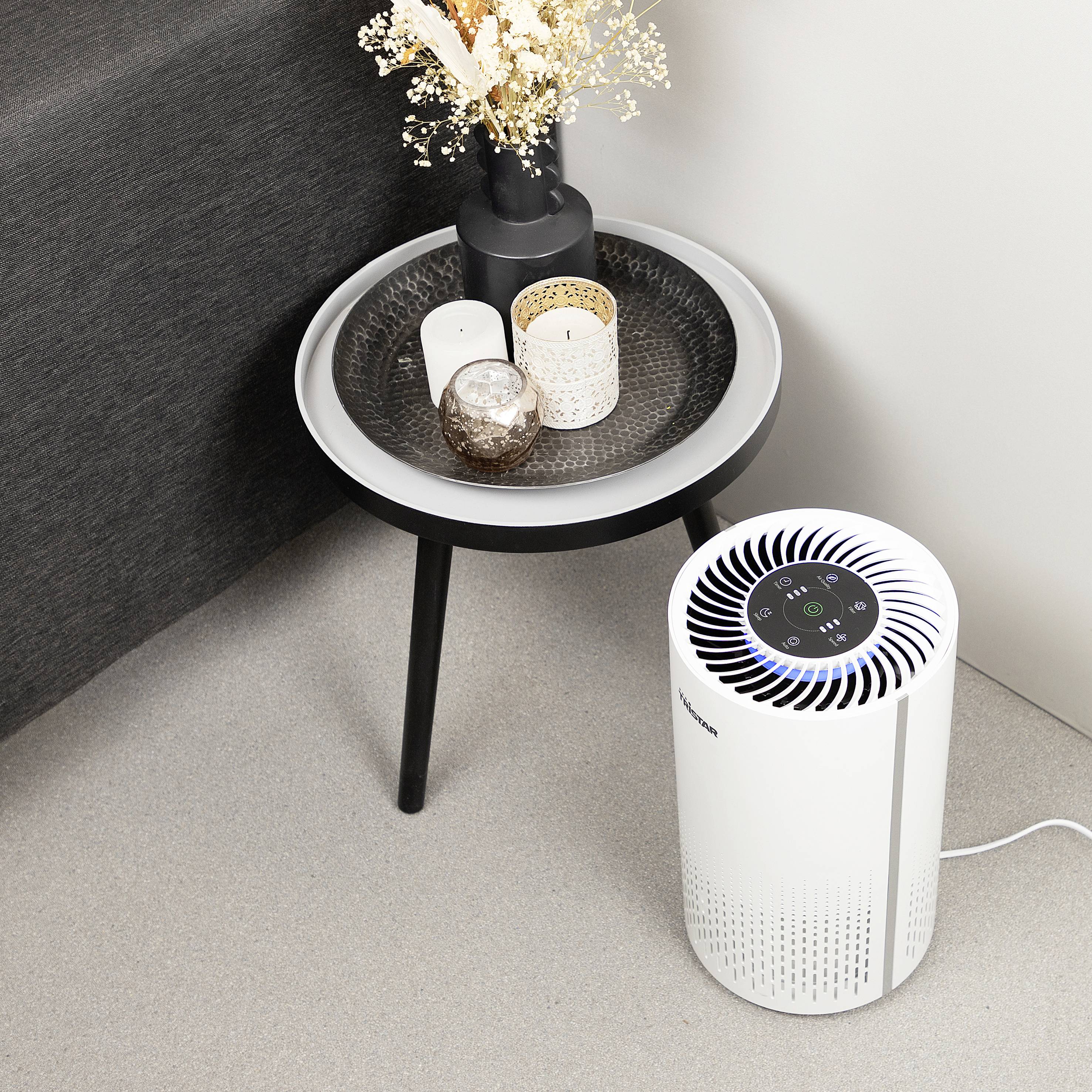 Un purificateur d'air est posé sur le sol à côté d'une petite table d'appoint avec des bougies et des fleurs séchées dans un vase.