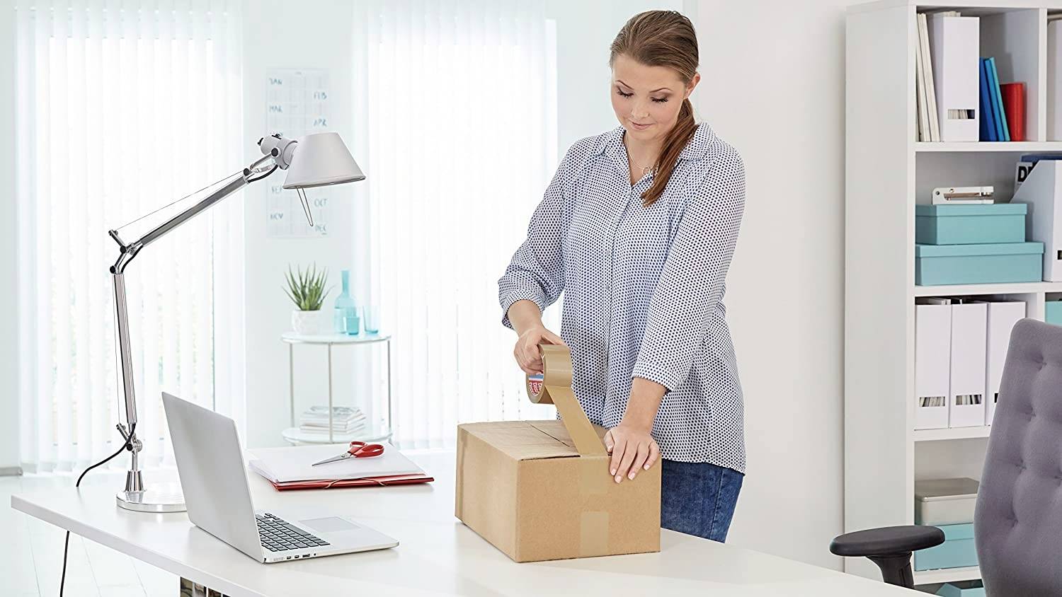 Une femme dans un bureau scelle un carton avec du ruban adhésif. Un bureau avec une lampe de bureau, un ordinateur portable et des dossiers en arrière-plan.
